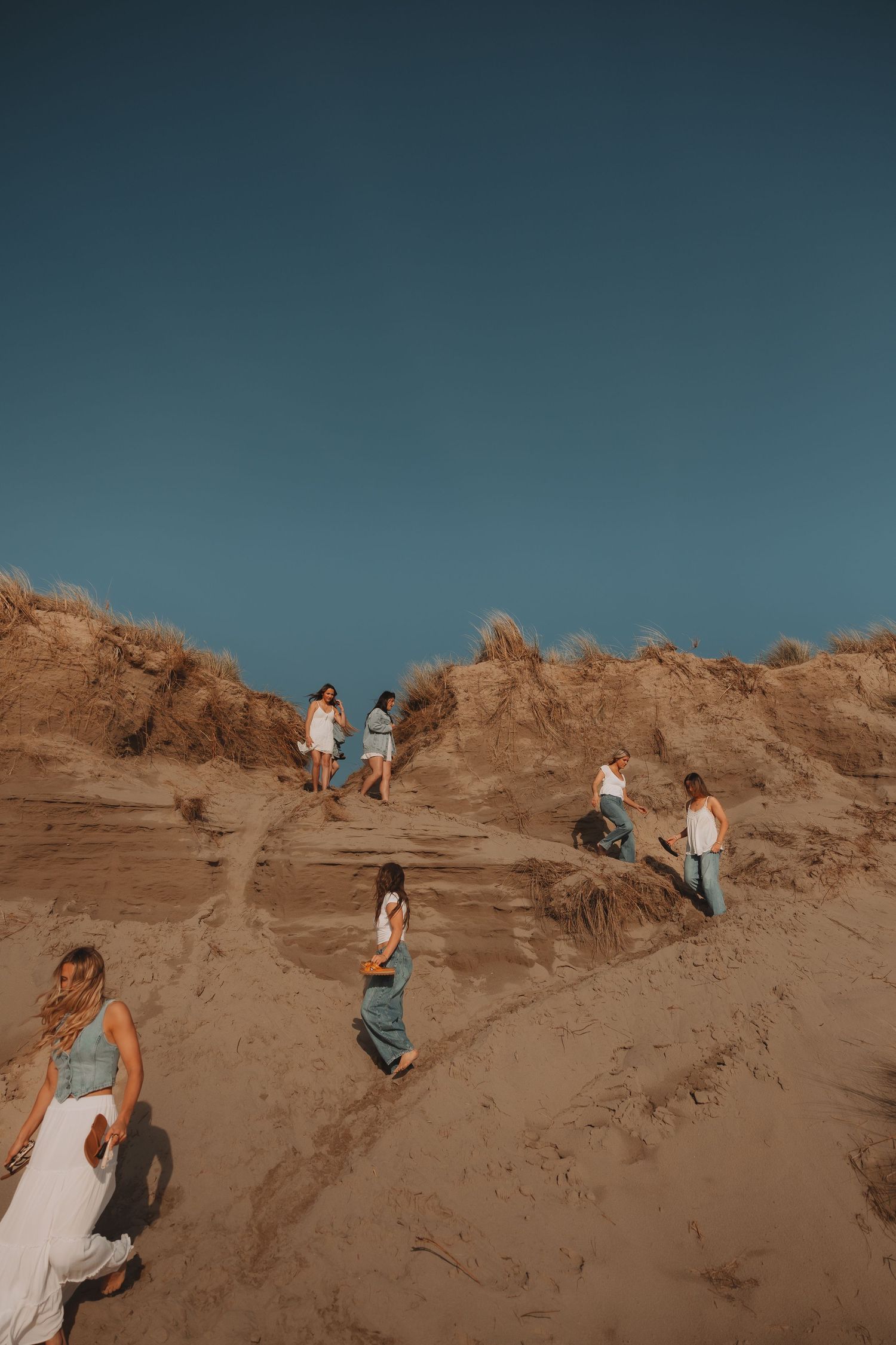 Group of young women climbing sandy beach dunes under a clear blue sky.
