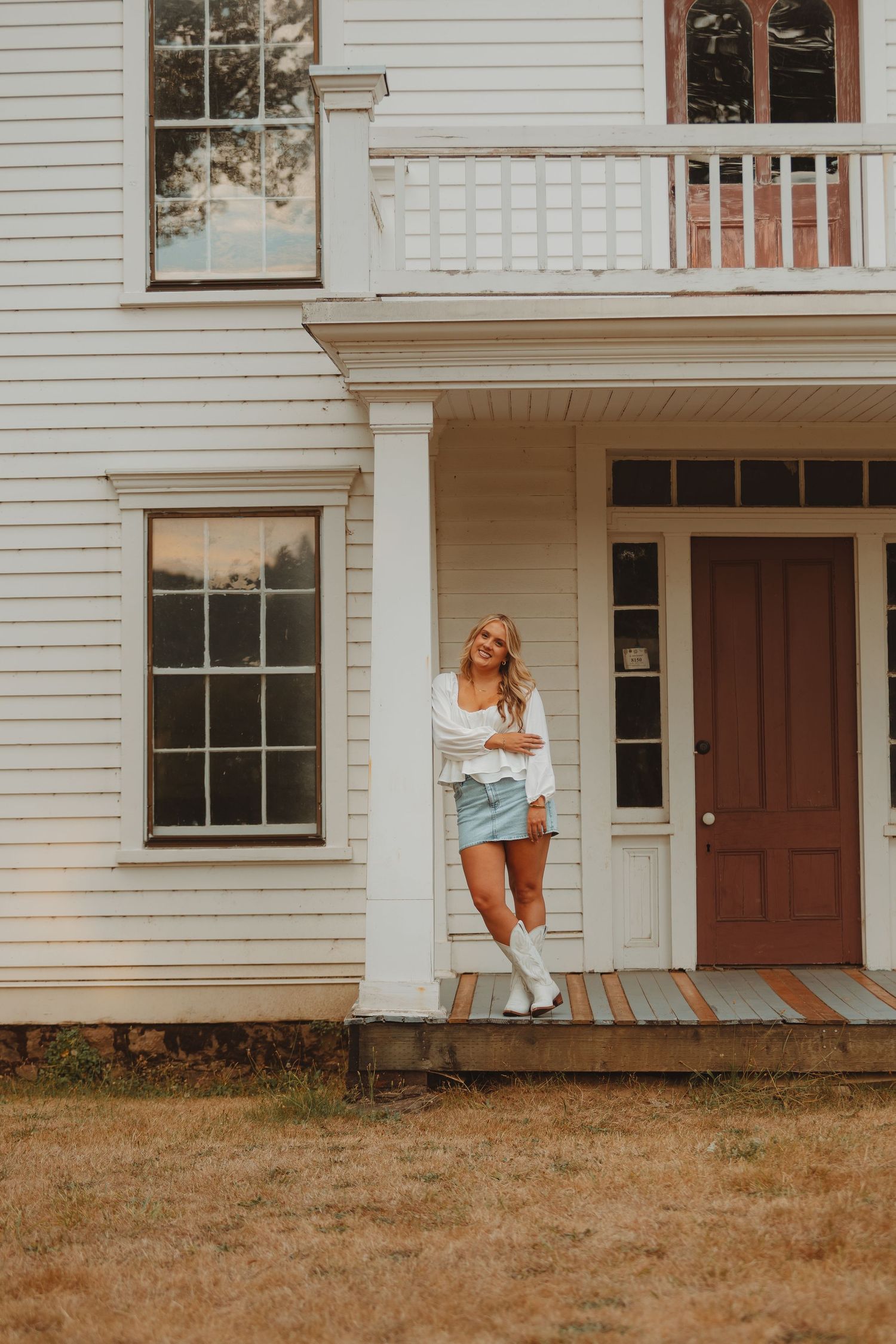 Smiling blonde woman in denim skirt and white boots posing by a classic white colonial house porch.