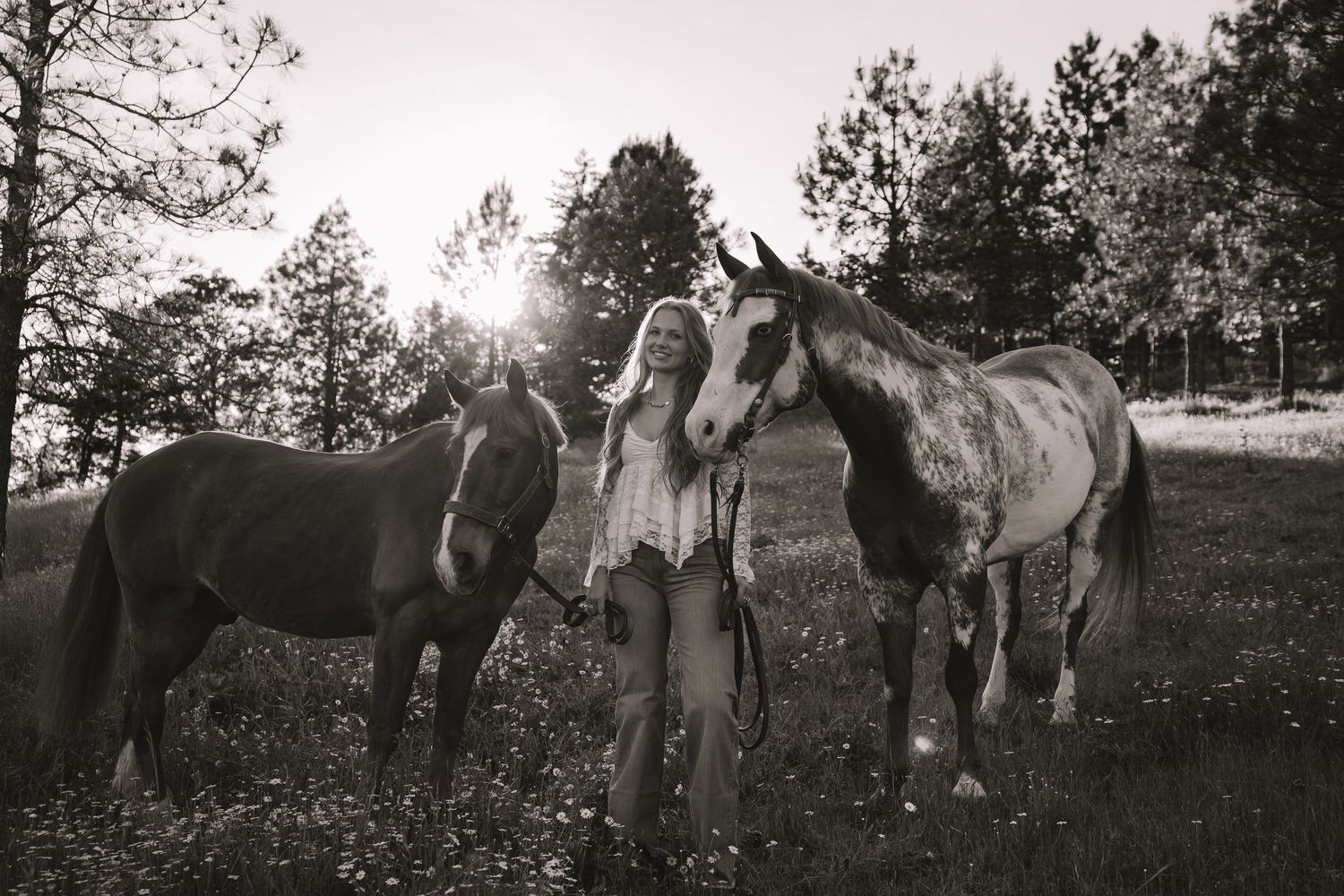 Black and white photo of a blonde woman standing between two horses in a sunlit field with trees in the background.