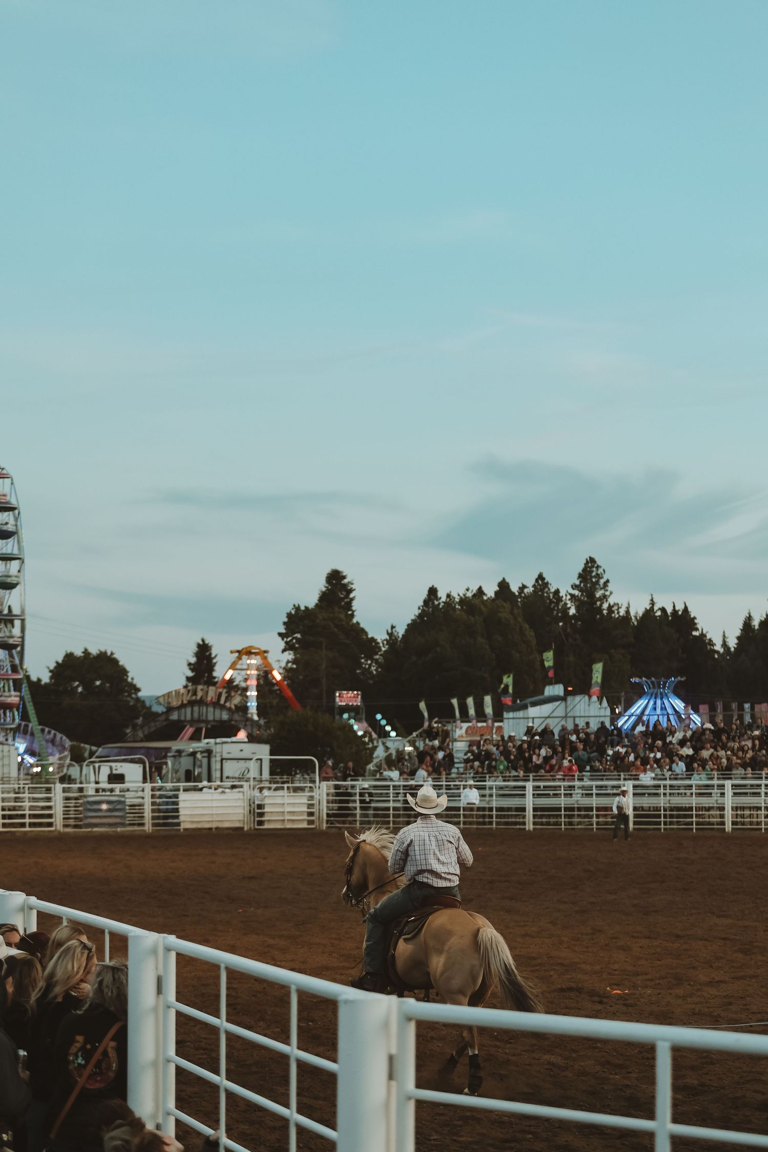 Rodeo scene with a crowded grandstand, ferris wheel, and rider in the dirt arena at dusk.