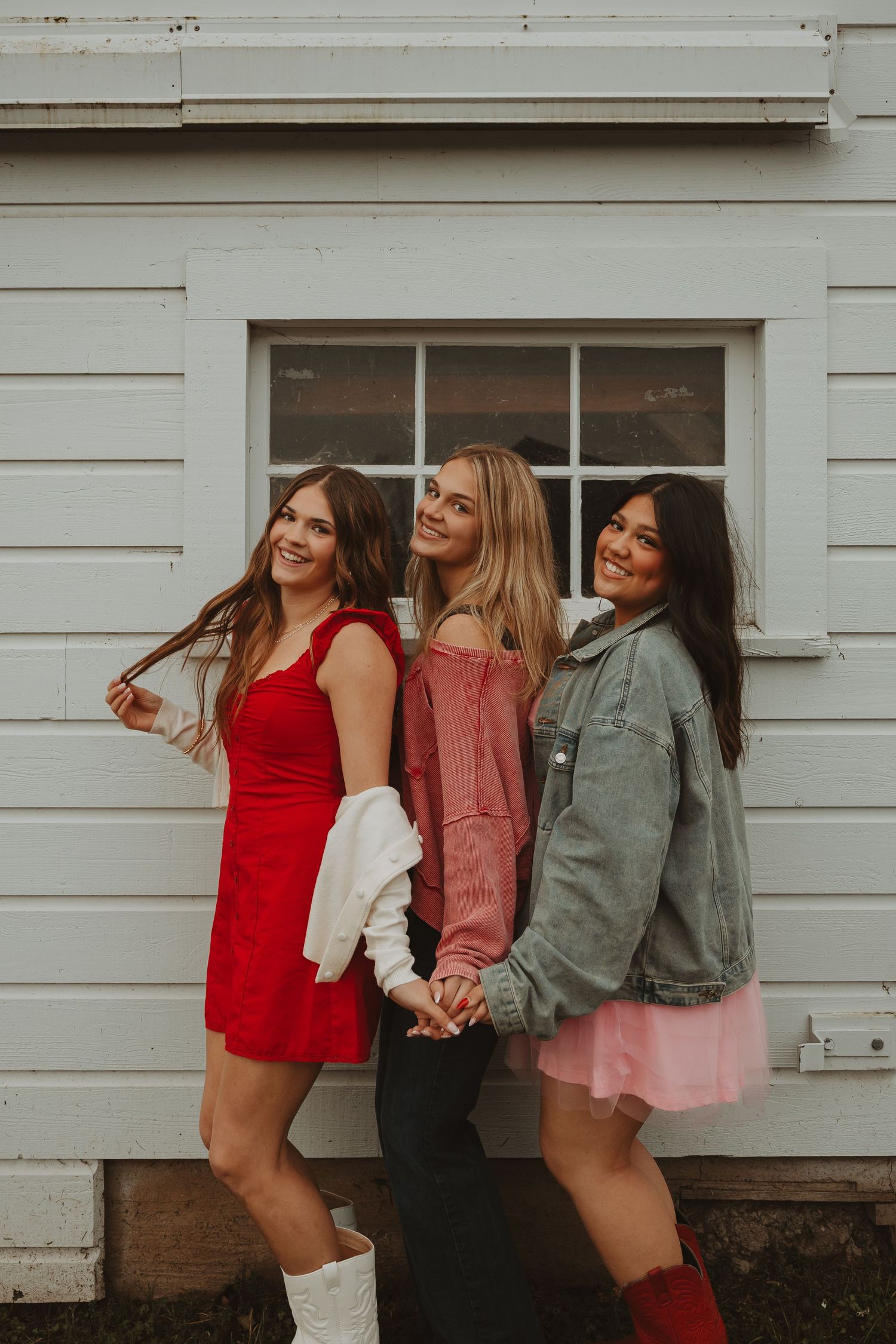 Three friends in red, pink and gray outfits pose playfully against white siding.