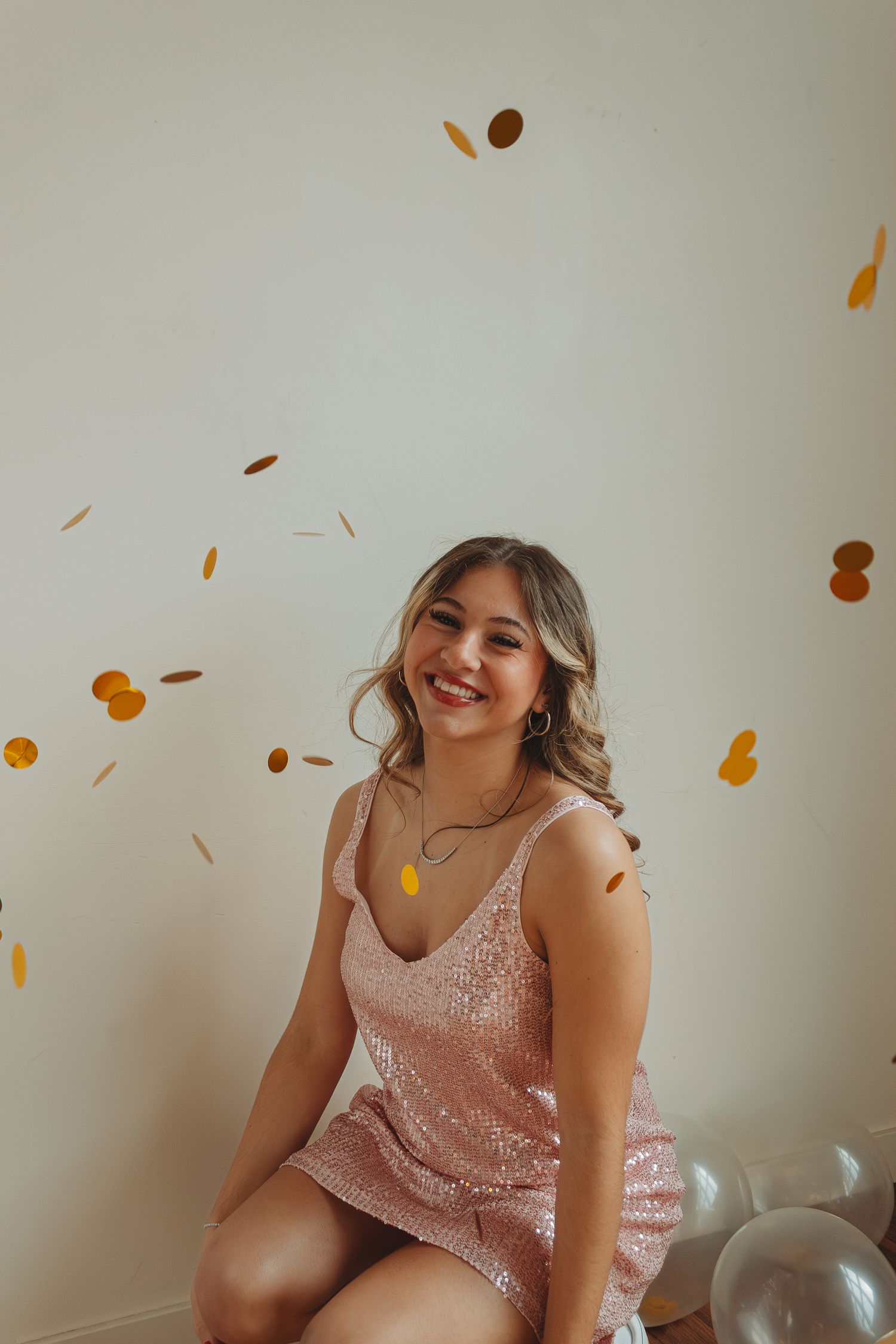 Person in pink sequin dress posing with confetti and balloons against white wall.