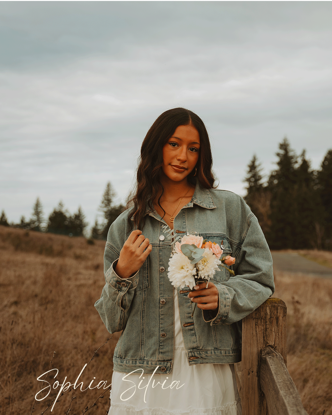 Individual in denim jacket holds delicate floral bouquet against scenic rural backdrop.