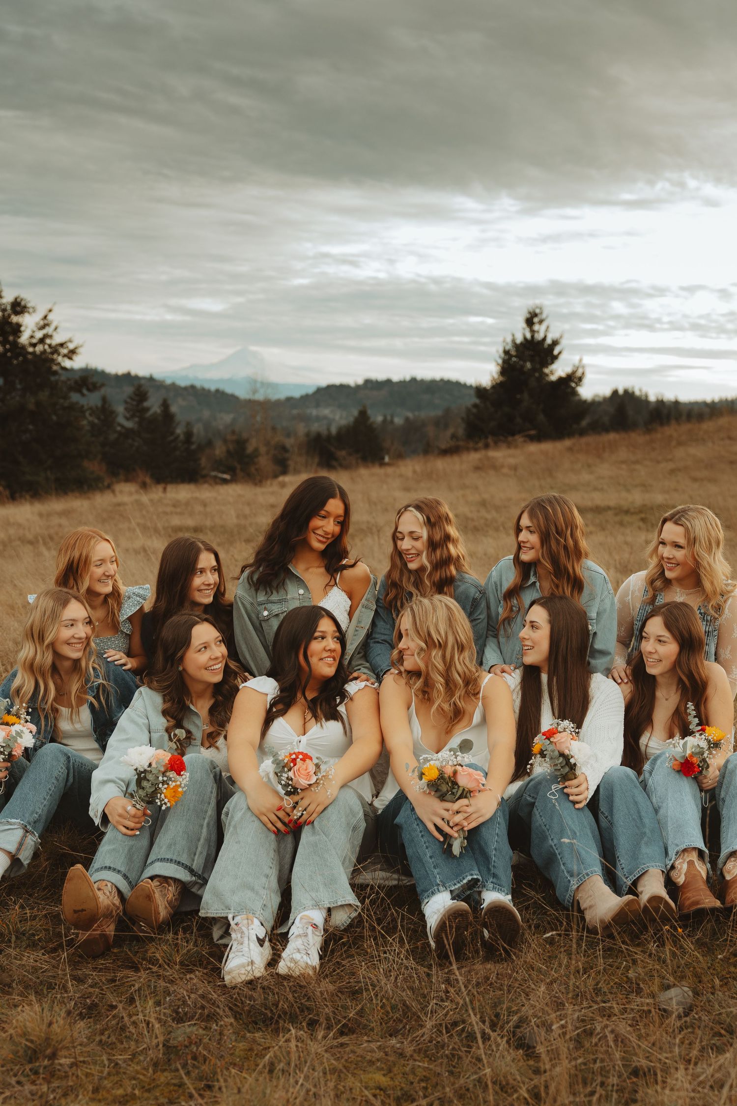 Friends in coordinated denim attire lay in a line on grass holding flowers with mountains in background.