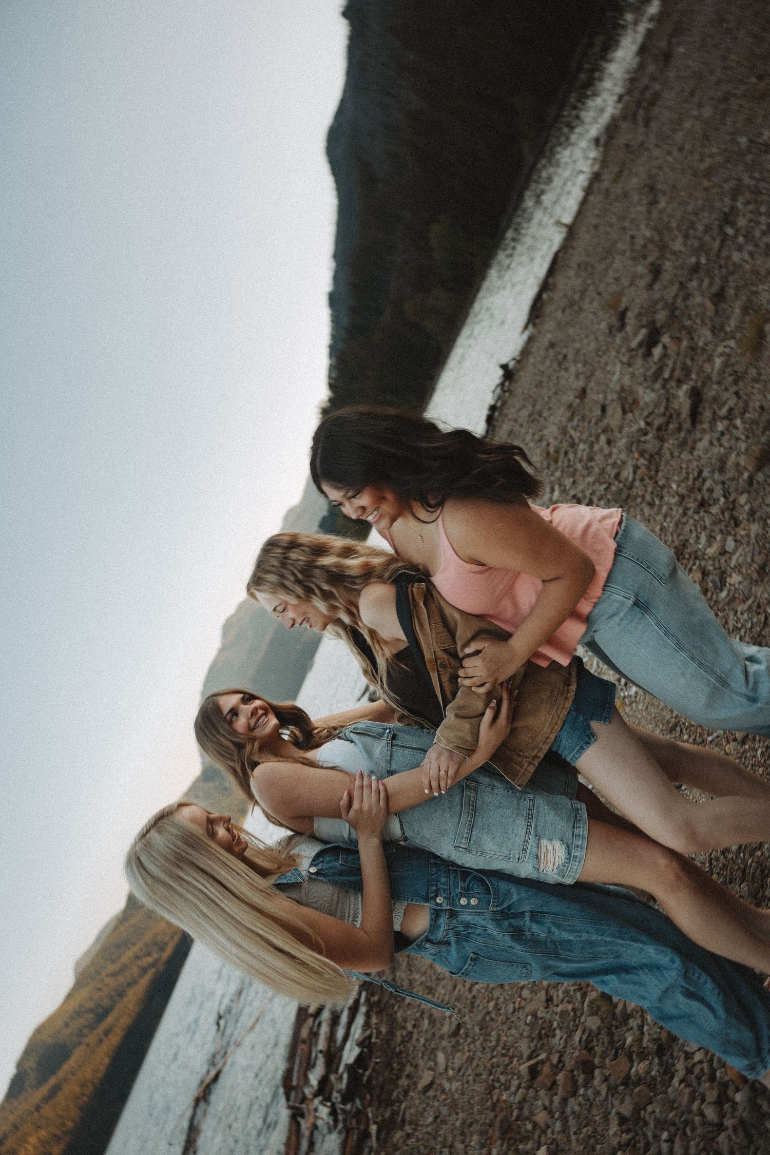 Four young women laughing and sitting together on a rocky beach at sunset.