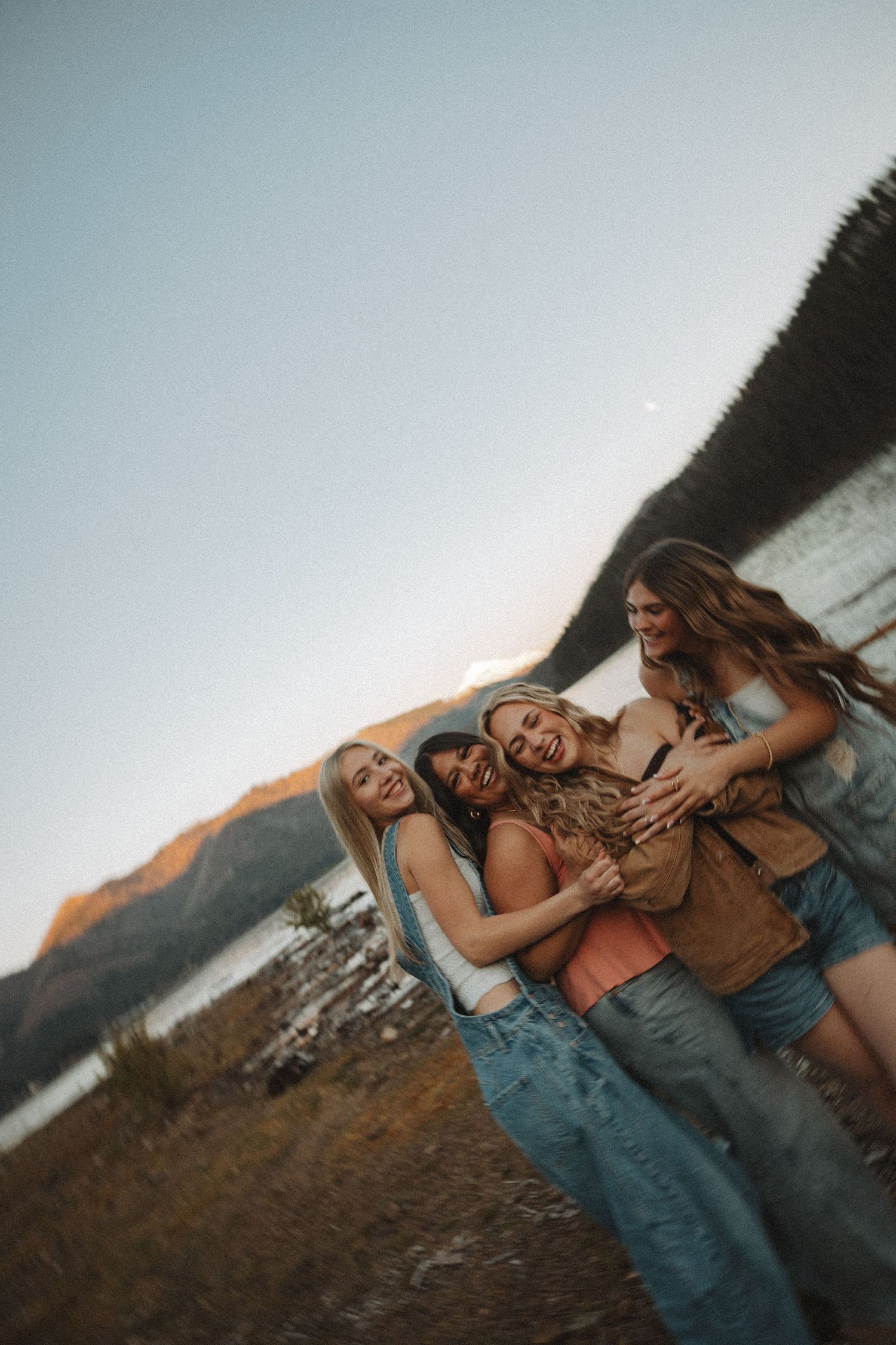 Three young women laughing and hugging by a mountain lake at golden hour.