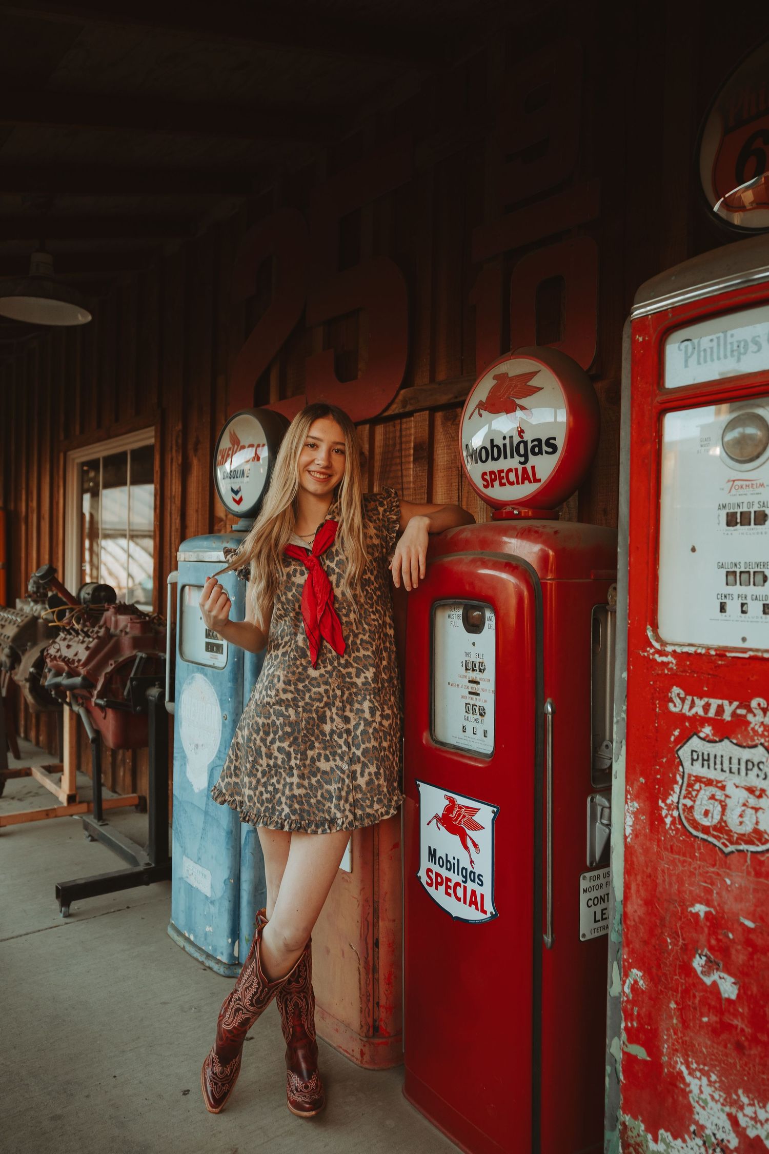 Young woman in leopard dress and cowboy boots leaning on vintage Mobilgas Special red gas pump indoors.