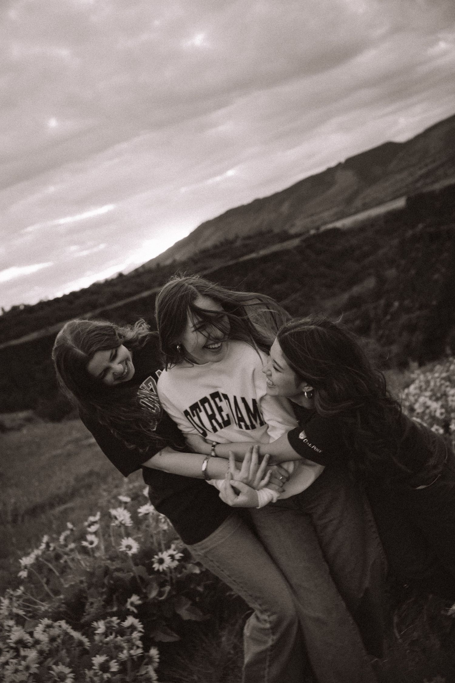 Three young women laughing and hugging in a flower field with mountain backdrop in black and white.