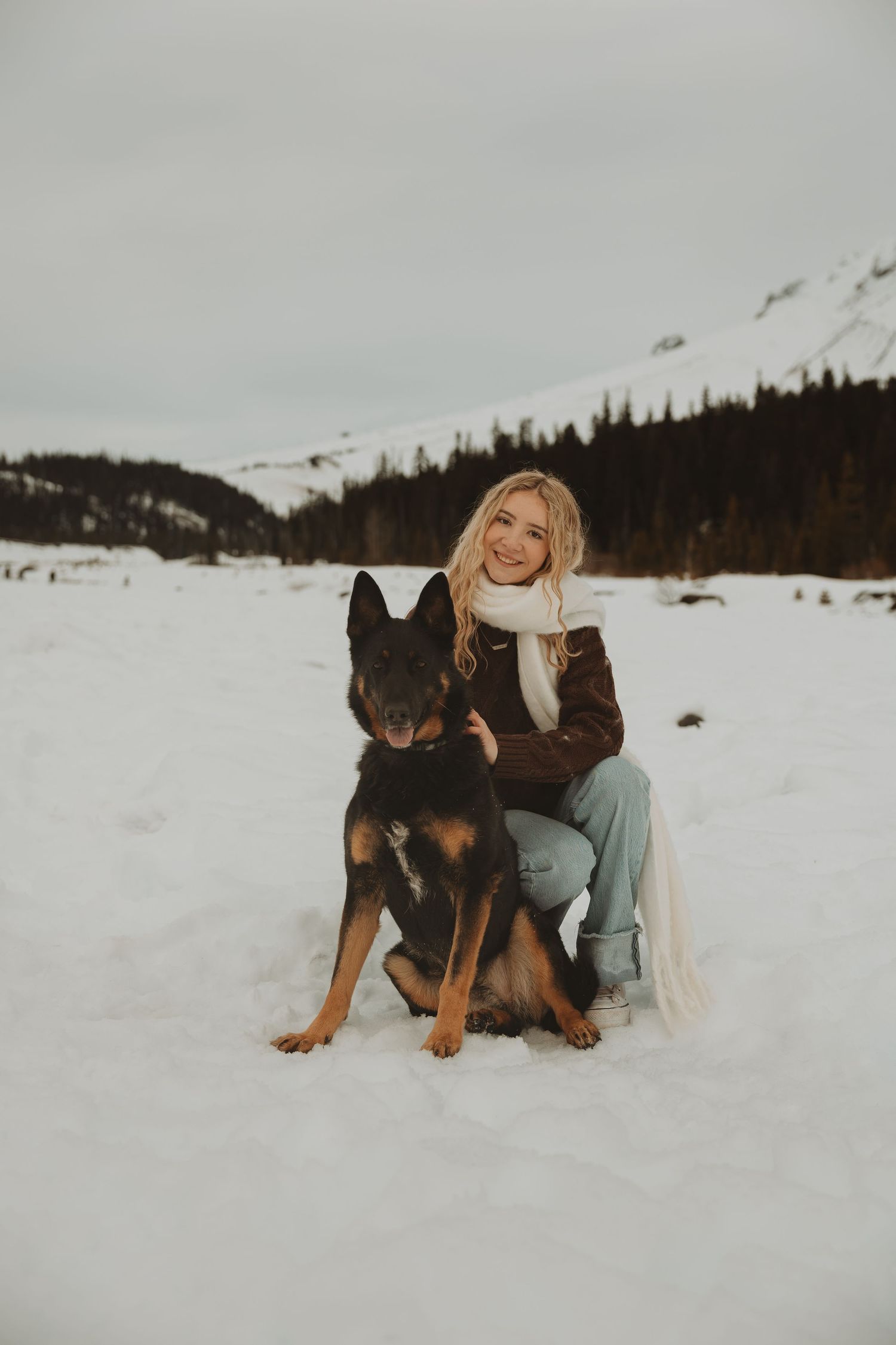Smiling blonde woman in brown jacket and white scarf posing with German Shepherd dog in snowy mountain landscape.