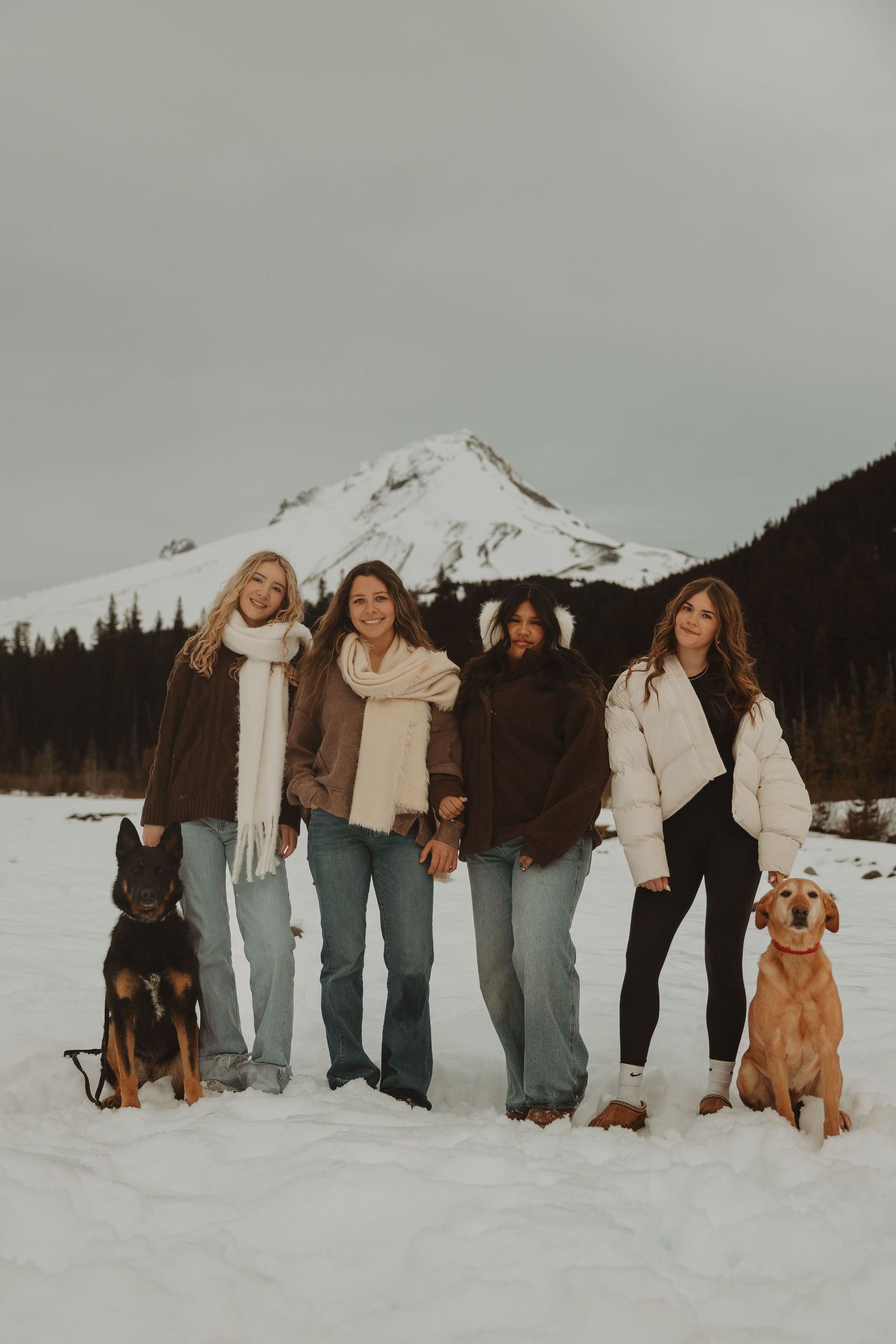 Four women with two dogs pose in front of snowy mountain peak in winter landscape.