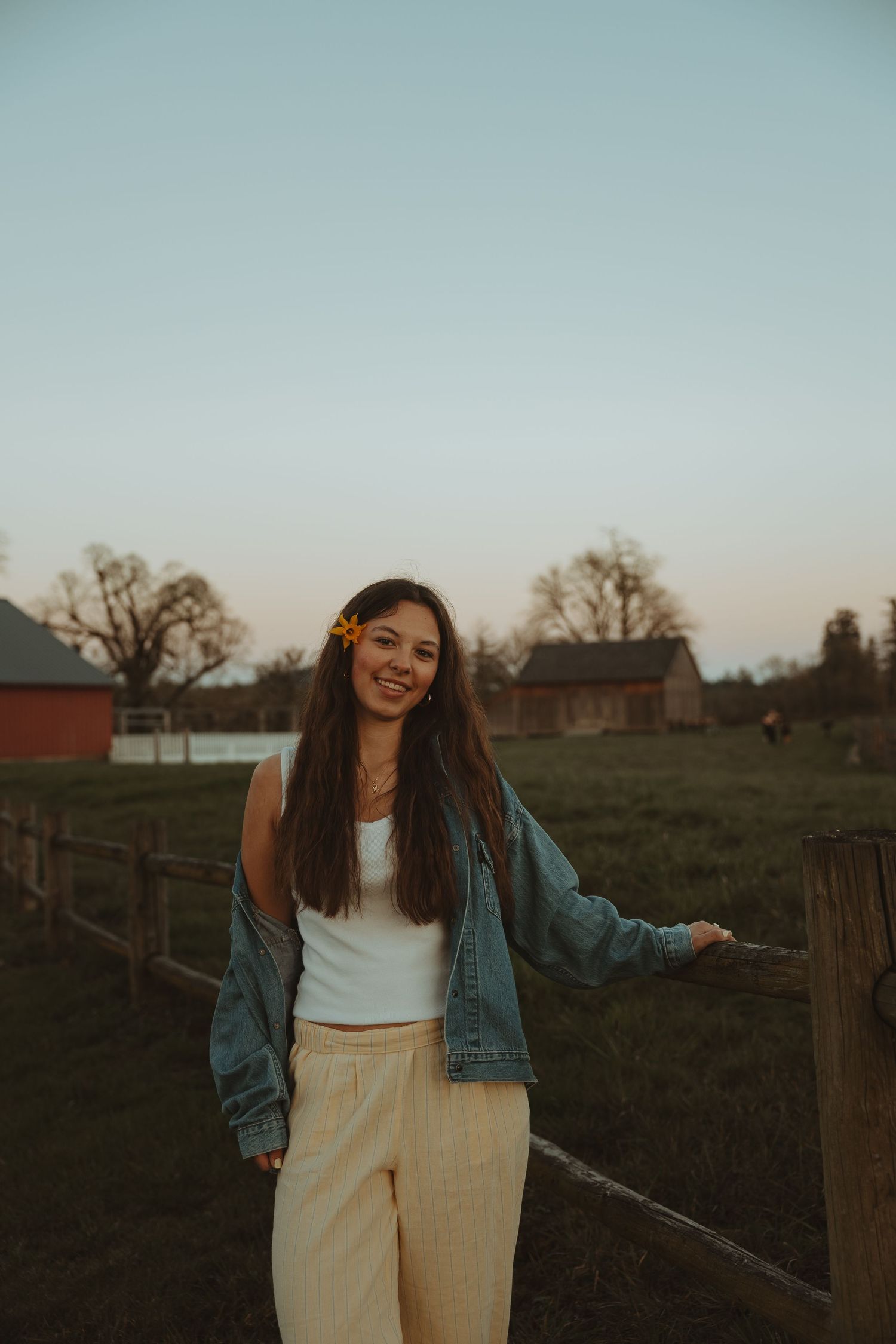 Happy young woman in denim jacket with yellow flower in hair stands by wooden fence at sunset farm.
