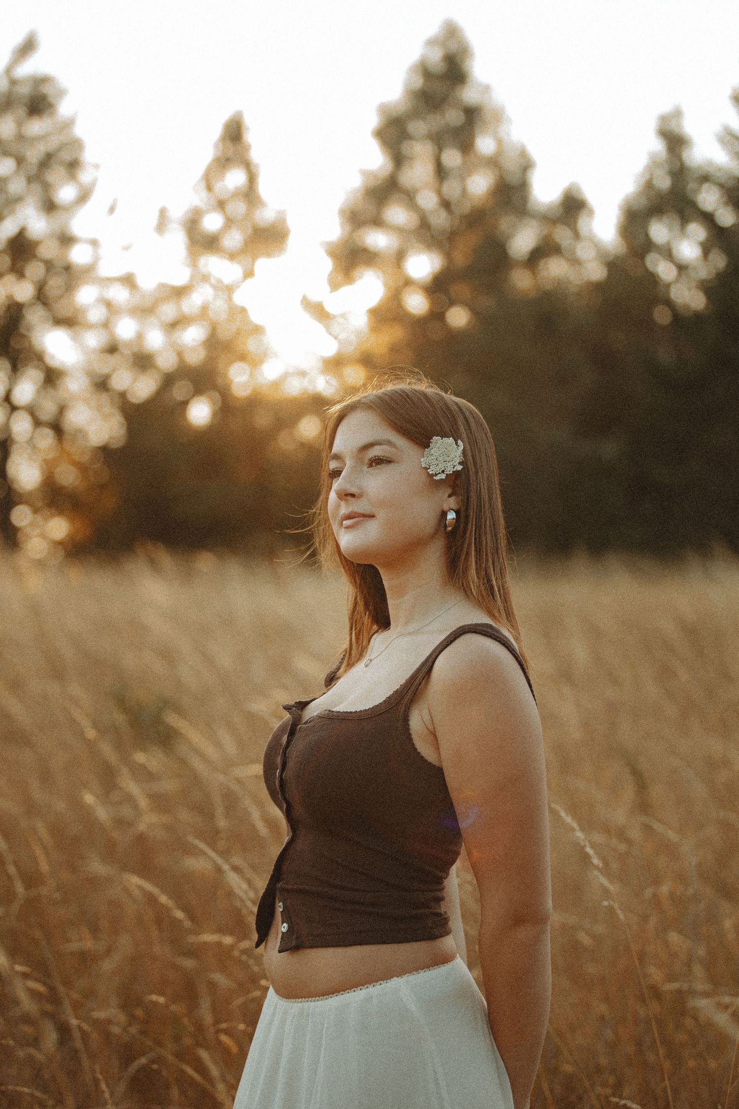 Young woman with flower hair clip in brown crop top and white skirt standing in golden field at sunset.