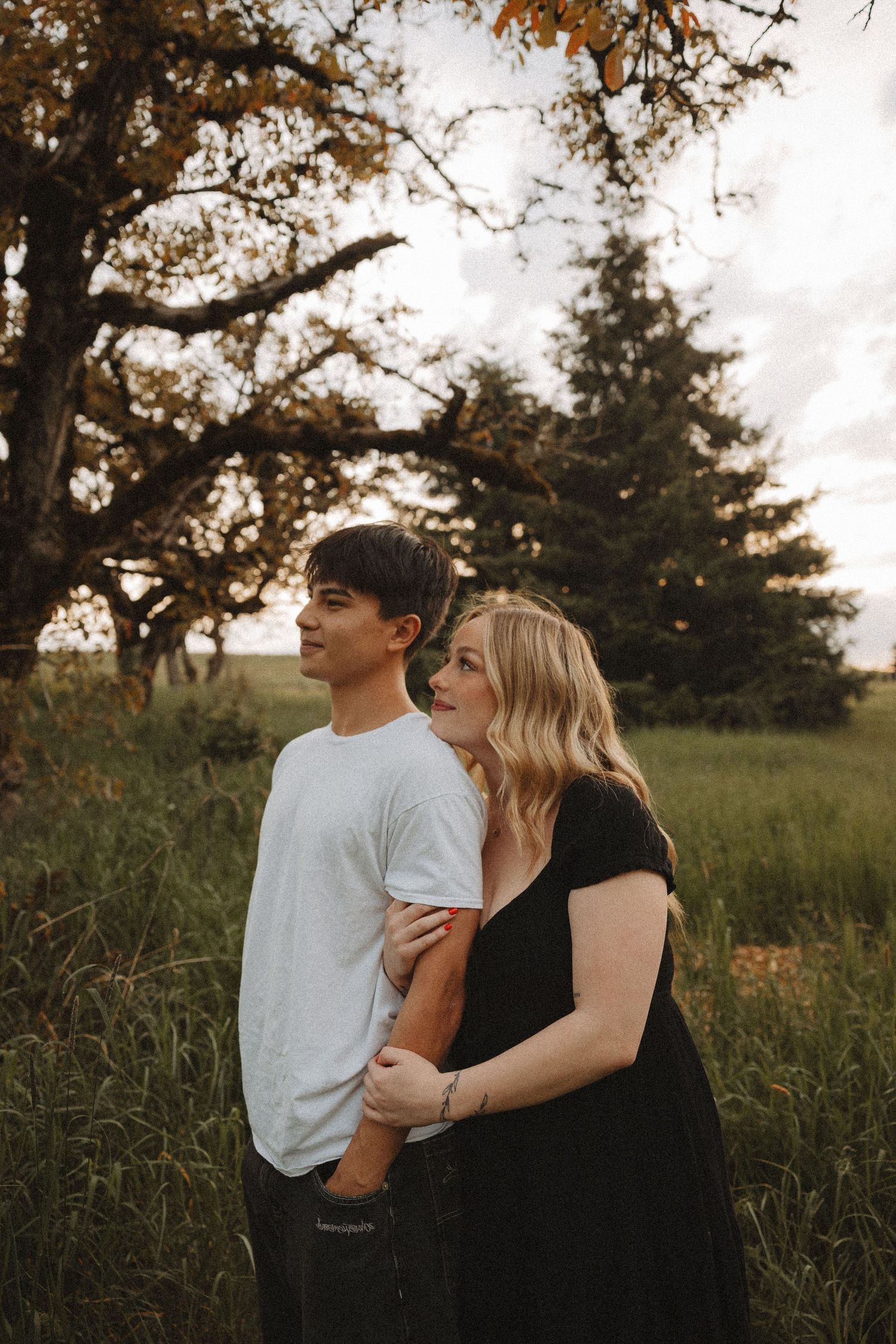 A couple embraces in a lush green field with golden autumn trees under a cloudy sky.