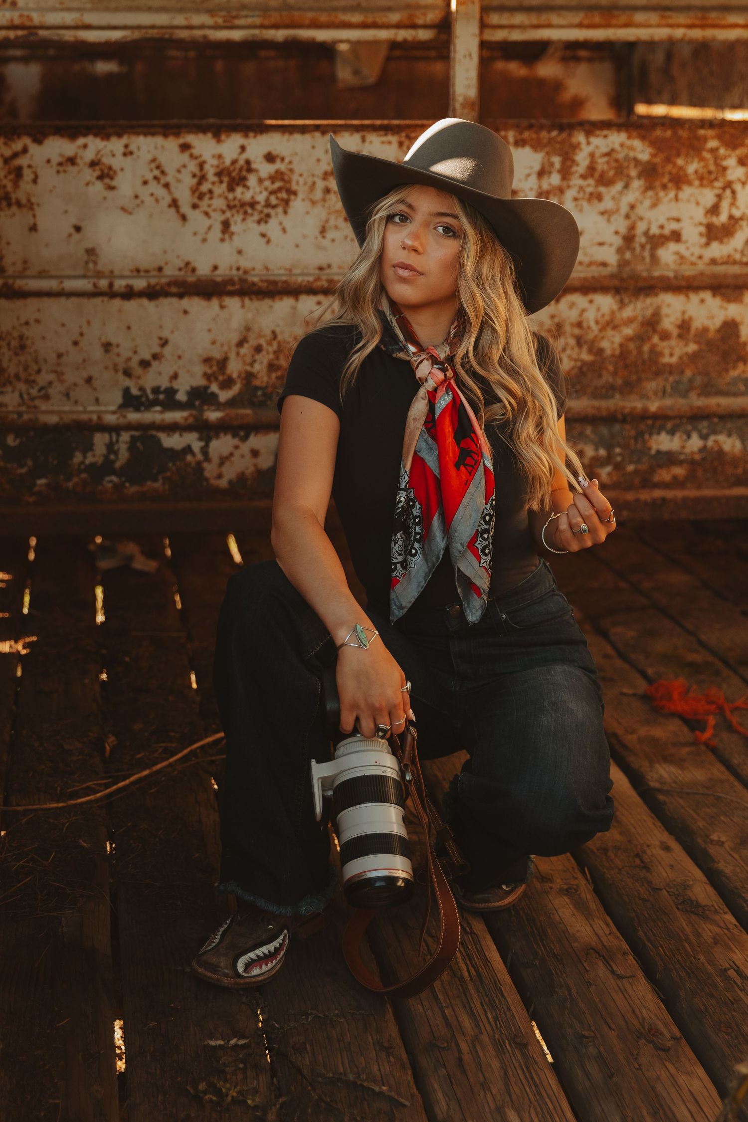 Western-styled photoshoot on rustic wooden floor with cowboy hat, boots and red bandana accessories in moody lighting.