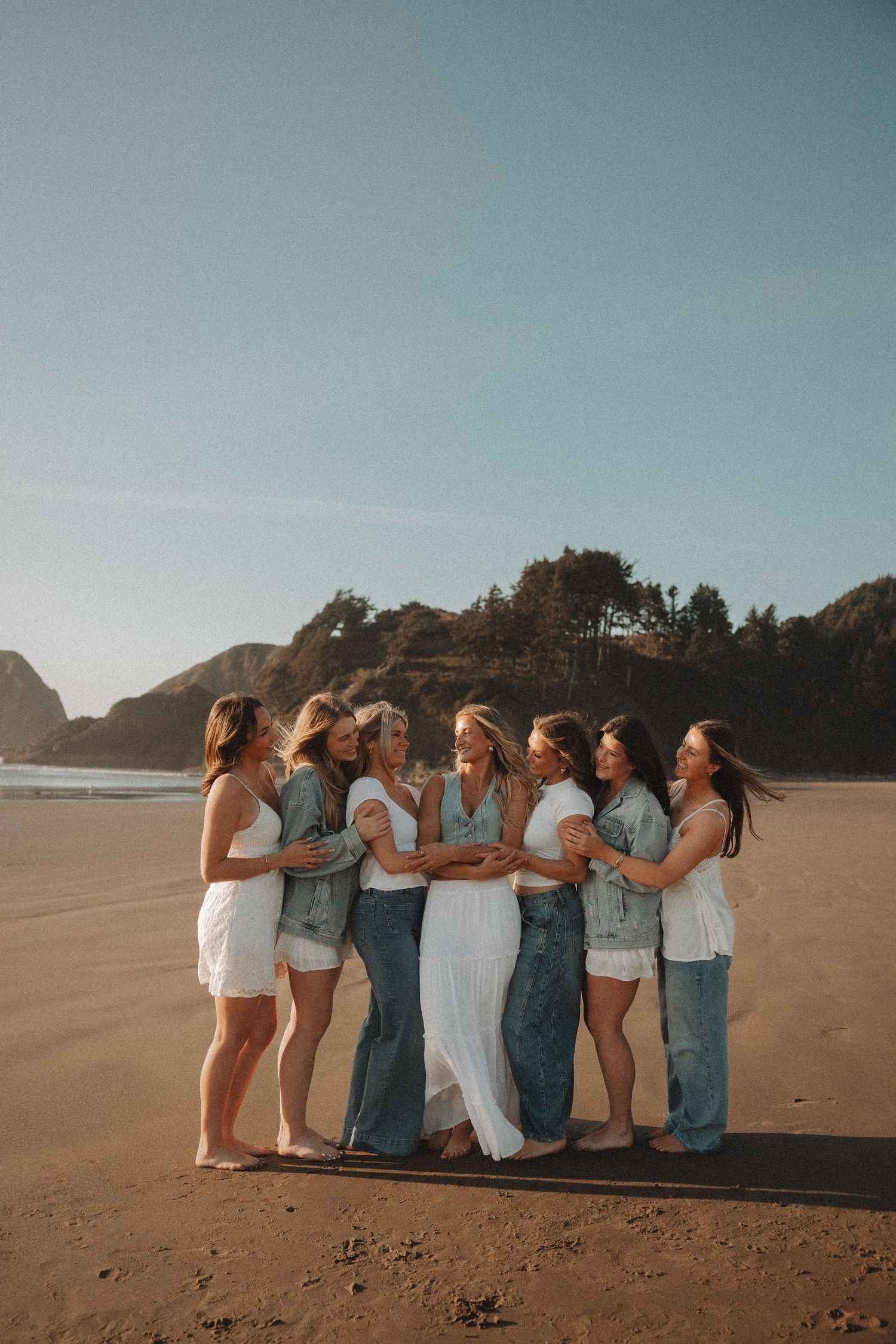 Eight women laughing and hugging on a beach at sunset, dressed in coordinating white and denim outfits.