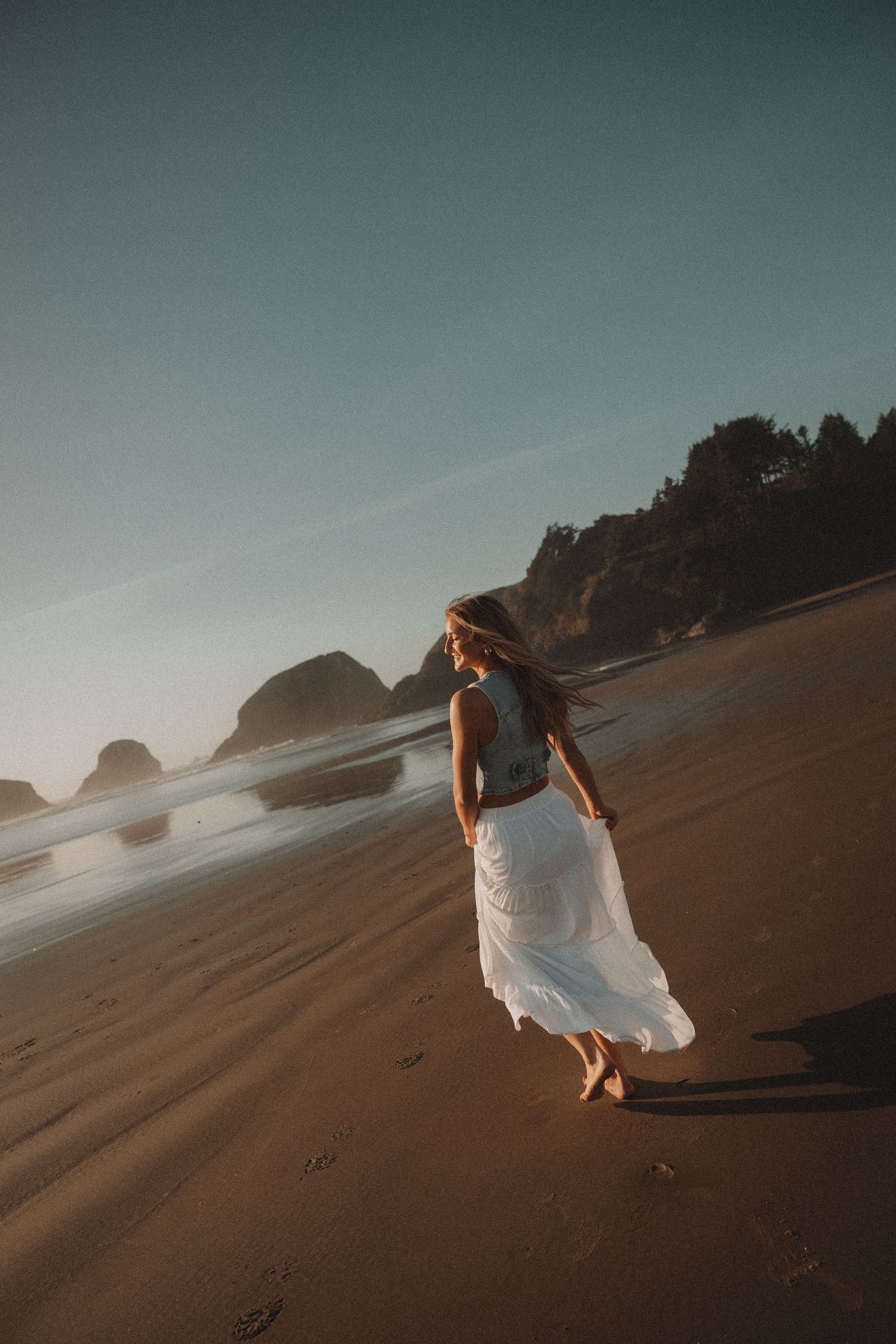 Woman in white dress walking on sandy beach at golden hour with rocky cliffs and ocean waves in background.