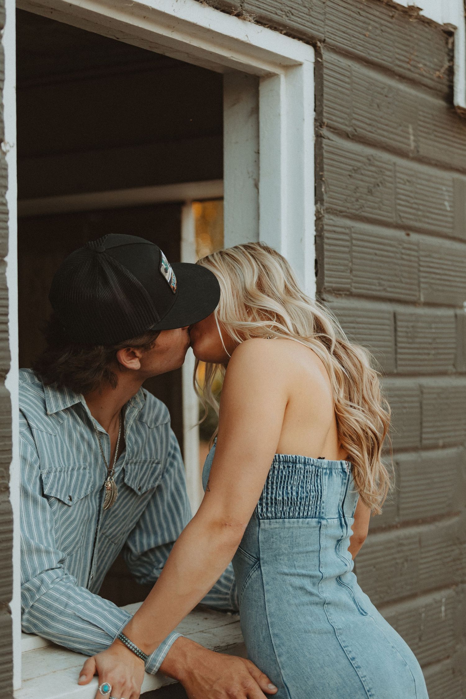 A couple shares a romantic kiss in a rustic doorway, the woman wearing a denim strapless outfit with long blonde hair.