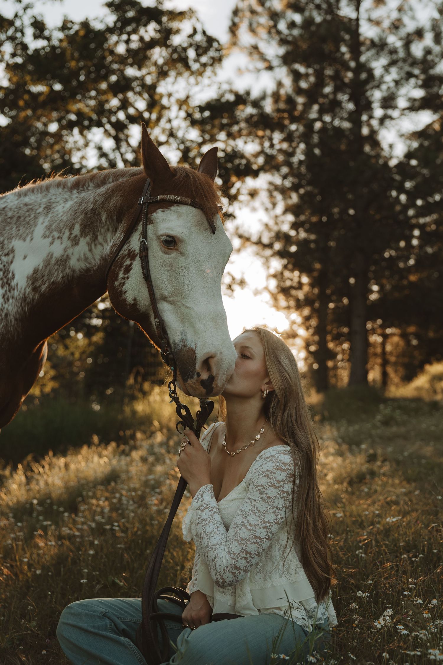 Young blonde woman kneeling to kiss a brown and white paint horse at golden sunset.
