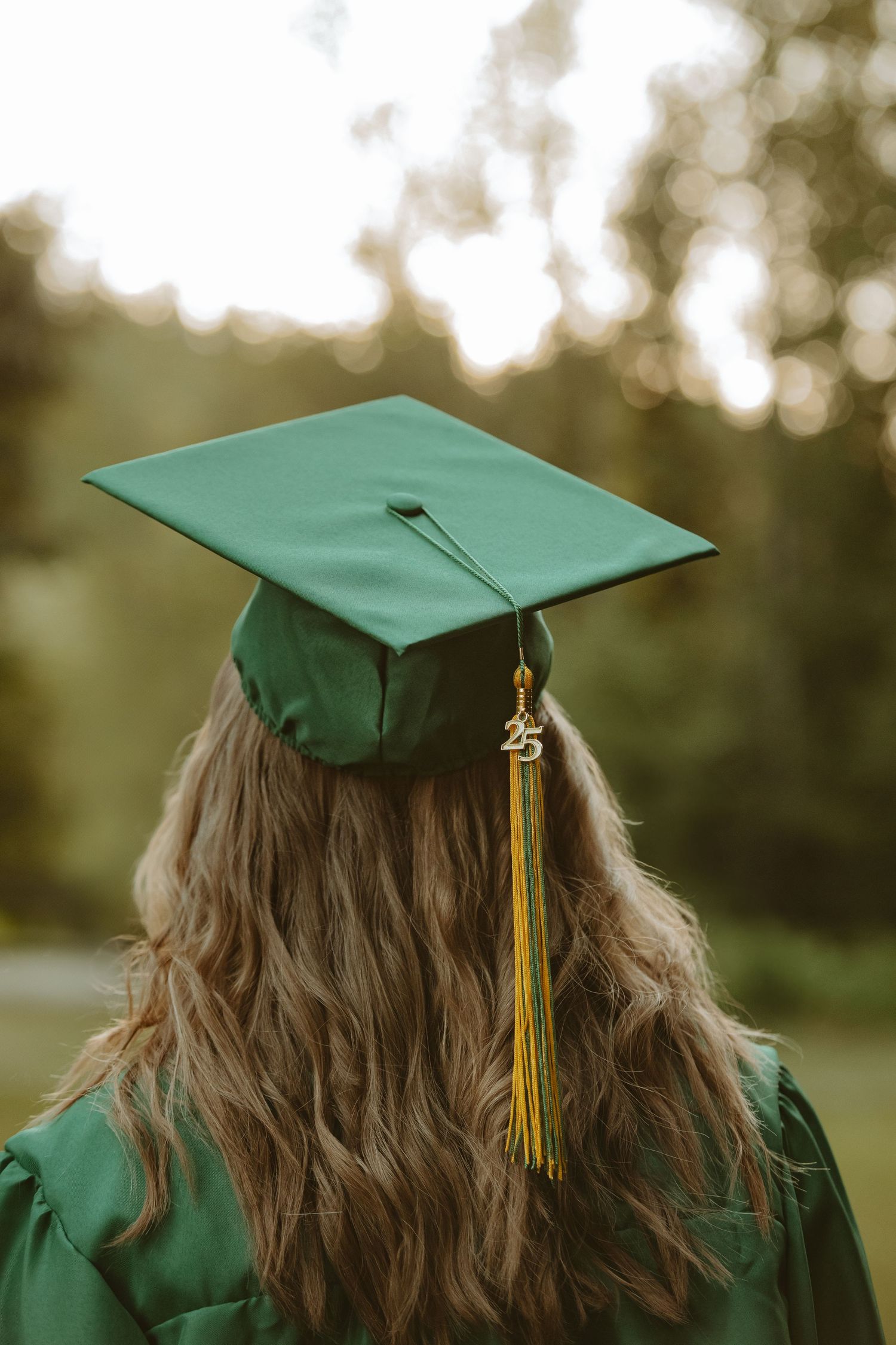 Back view of a graduate wearing green cap with gold tassel against blurred natural background.