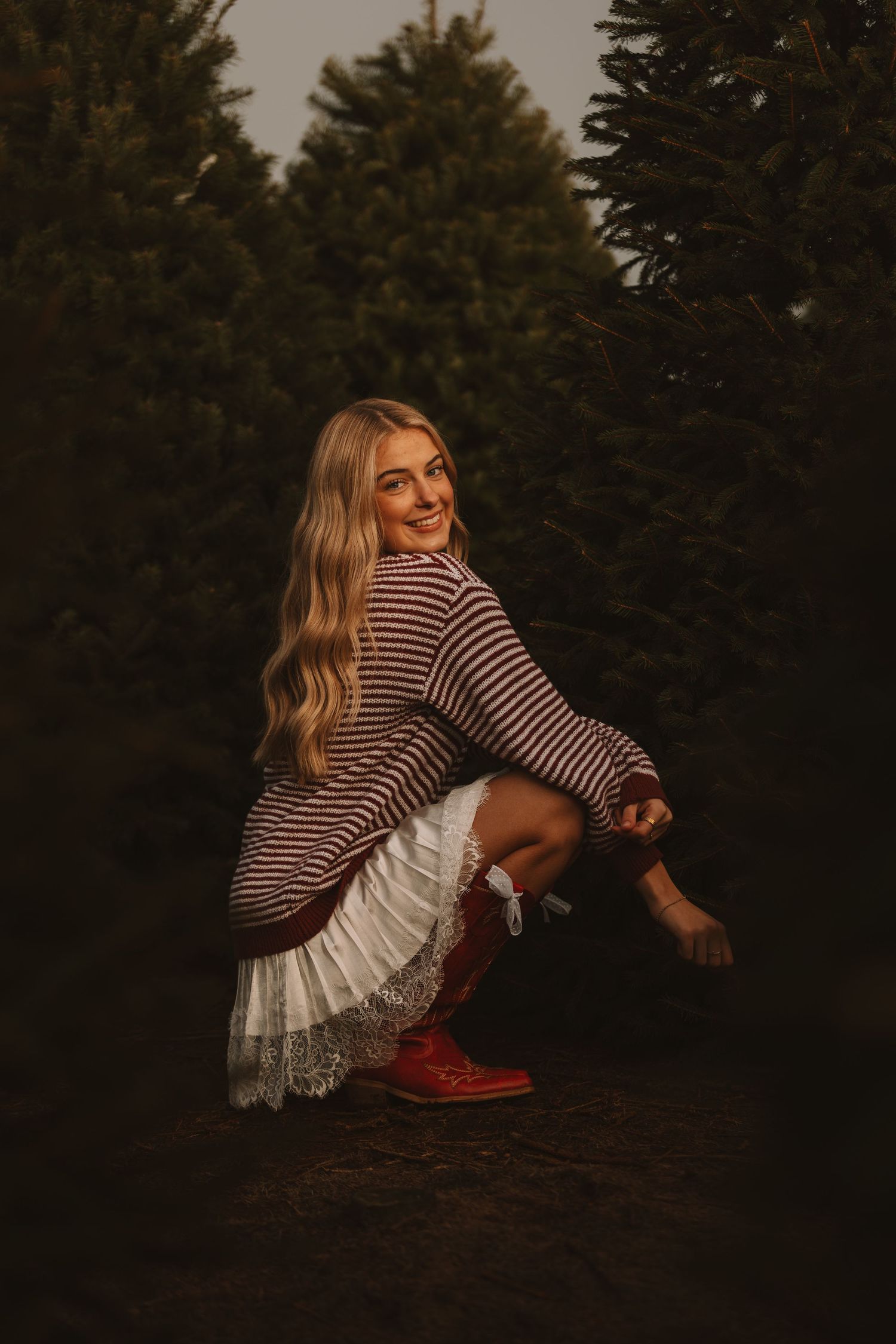 Person in striped top and white skirt poses artistically near pine trees at sunset.