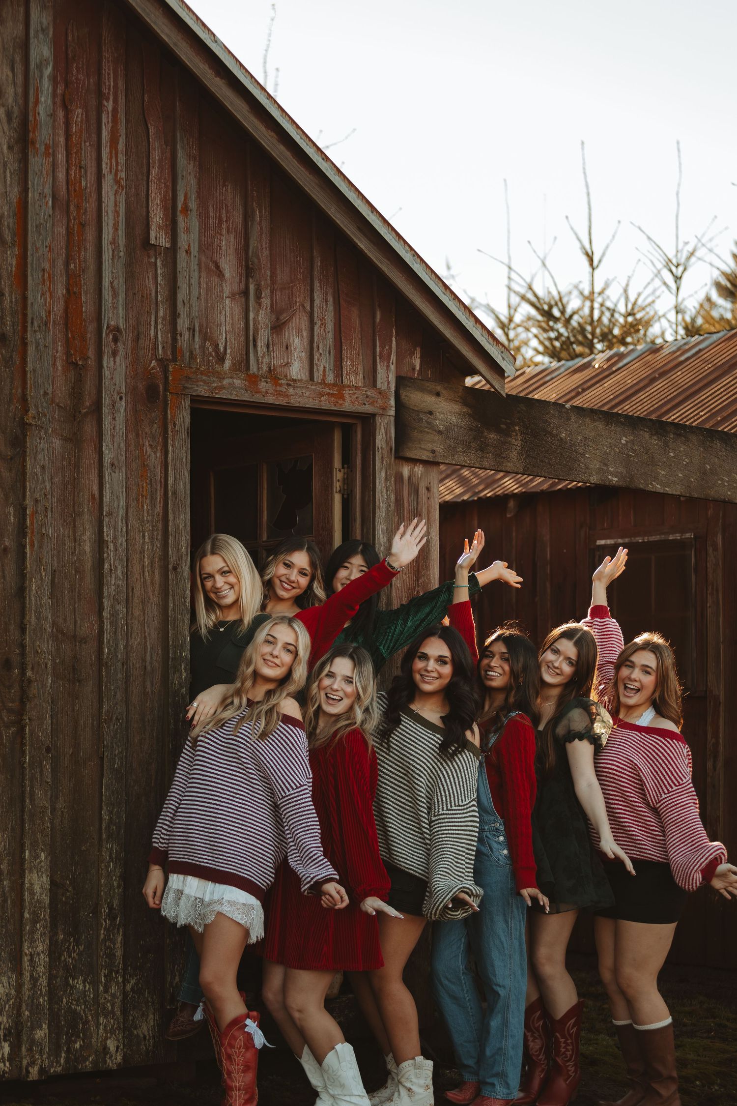 Friends in cozy sweaters and boots pose playfully against a rustic barn wall.