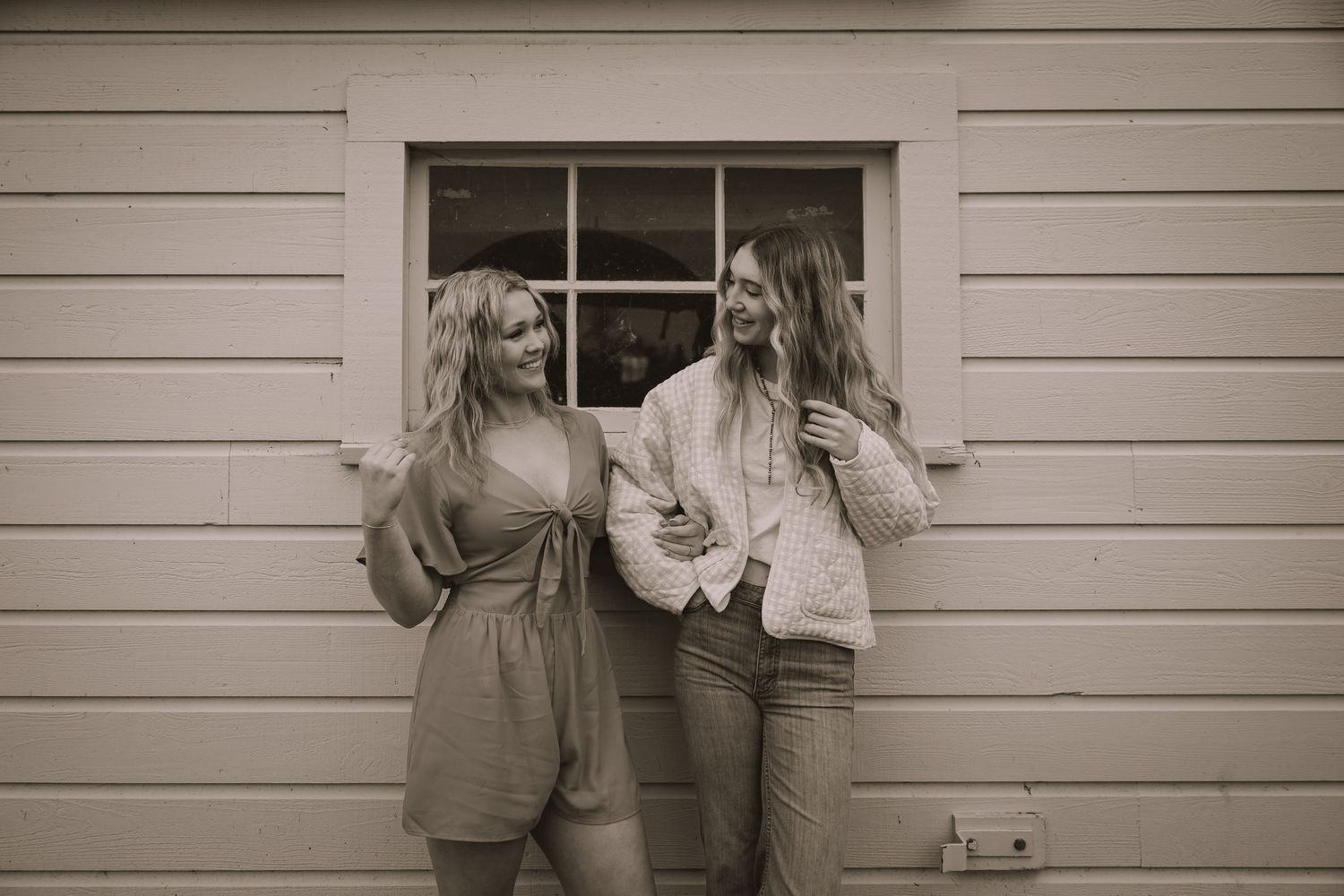 Two friends share a candid moment against white siding in a vintage-style black and white photo.