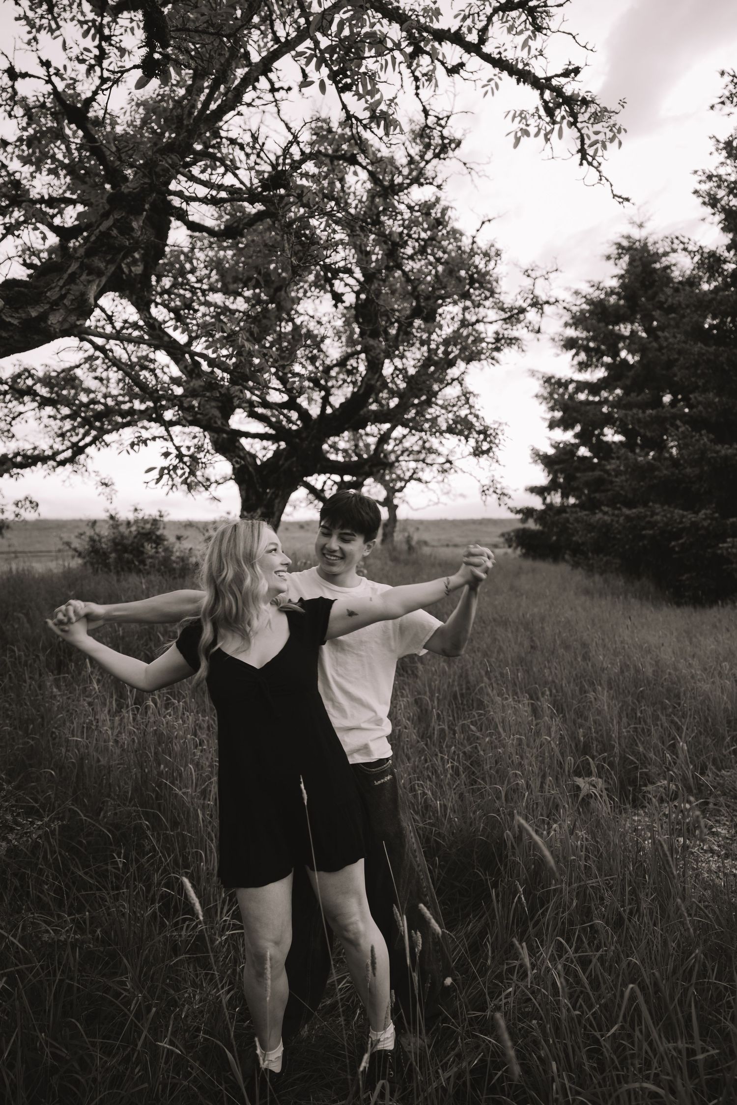 Black and white photo of a joyful couple dancing together in a field beneath a sprawling tree.
