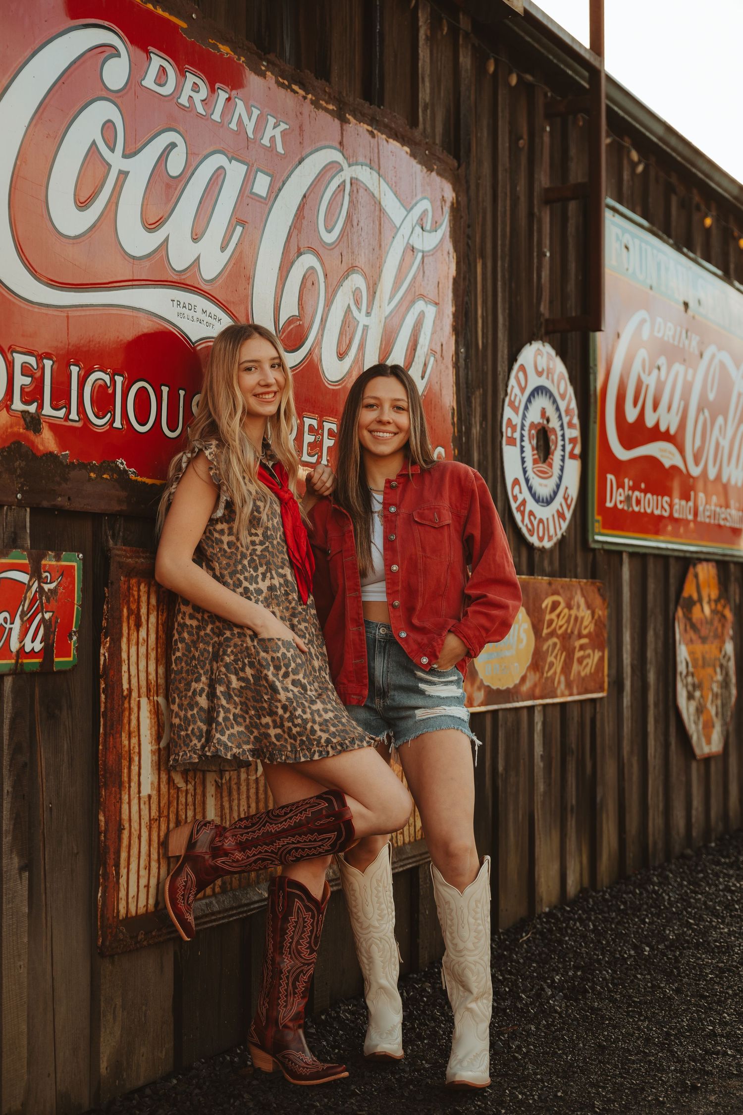 Two women in cowboy boots and western outfits posing against rustic wall with vintage Coca-Cola signs.