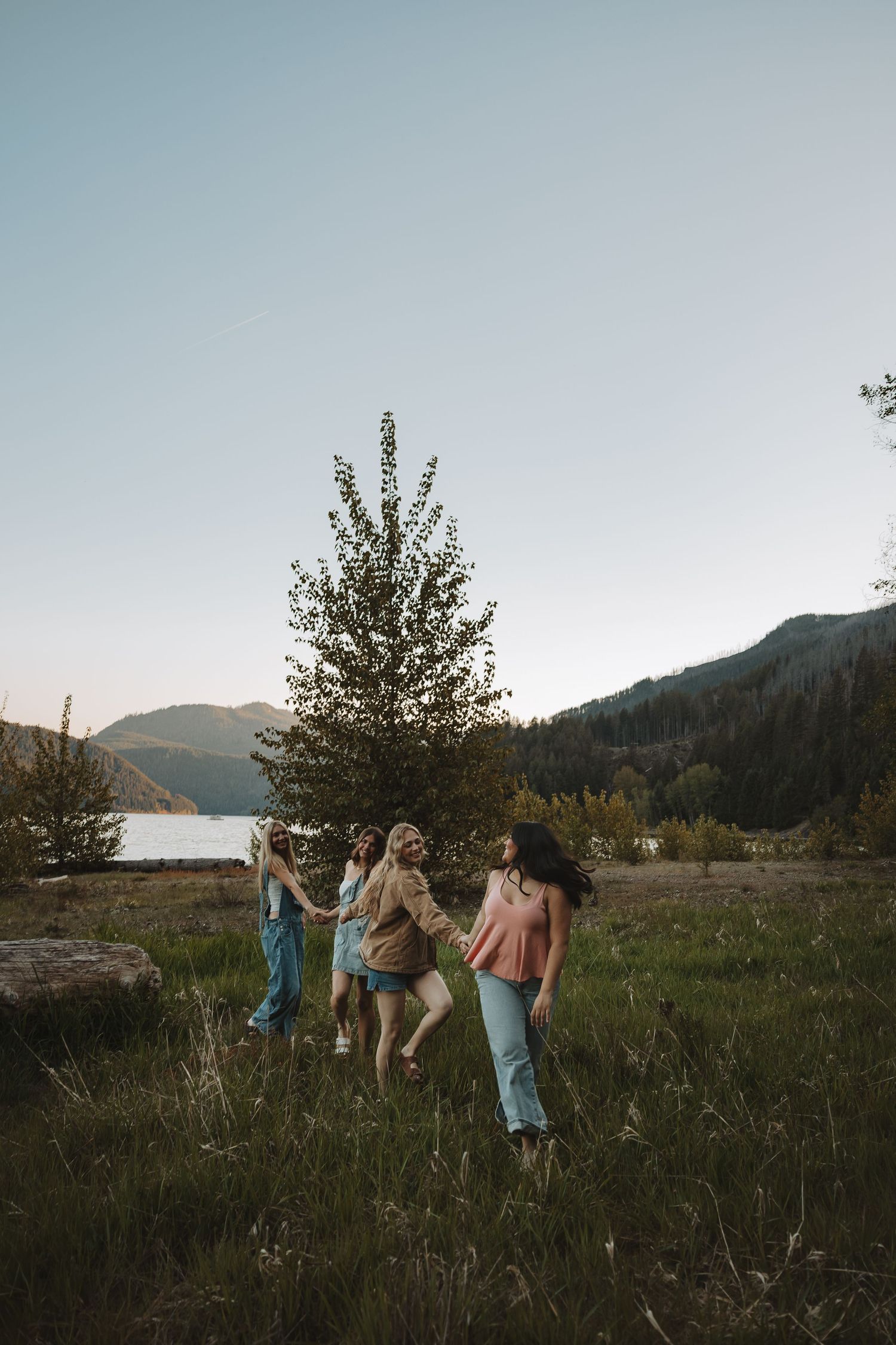 Four young women holding hands walking through a green meadow near a mountain lake at sunset.