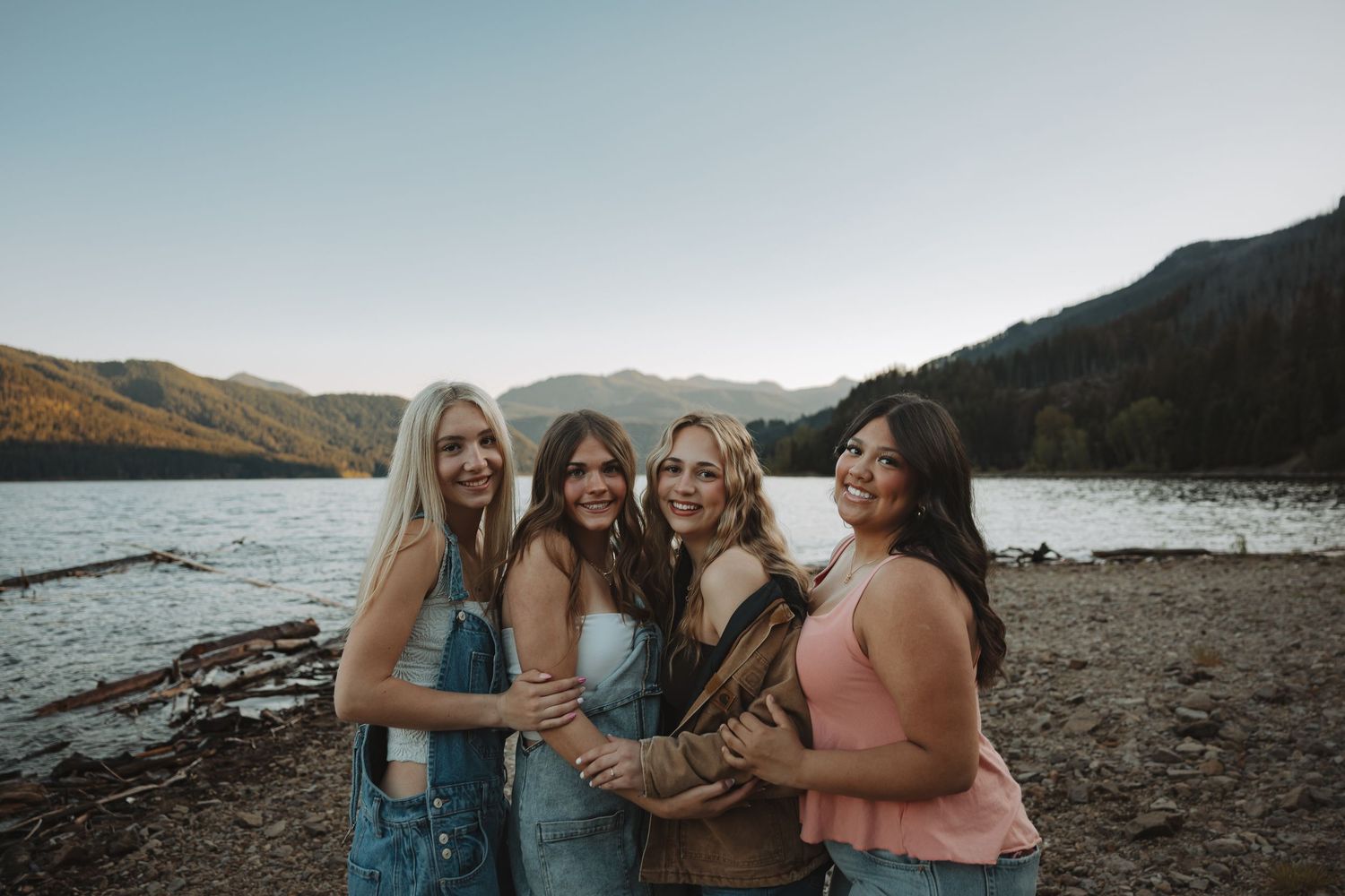 Four women smiling and hugging by a scenic mountain lake shore during golden hour.