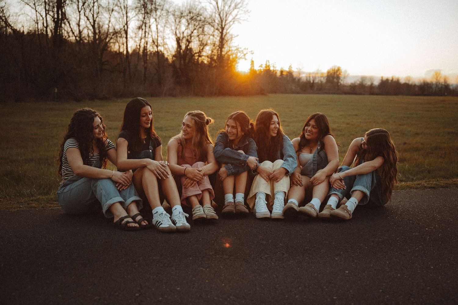 Seven girls sitting together on a road laughing at golden hour sunset in a park.