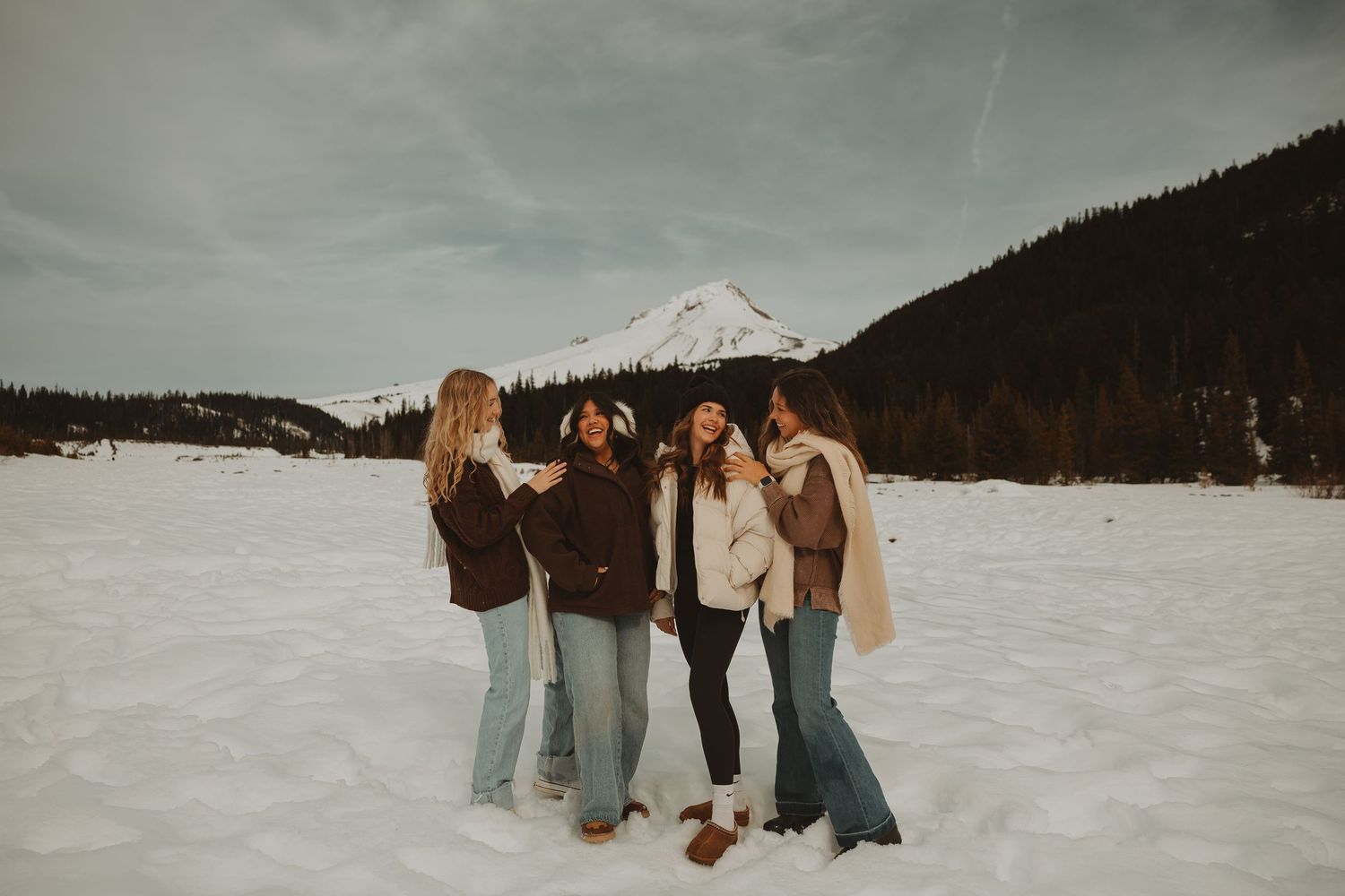Four women laughing together in a snowy mountain landscape with evergreen trees and a snow-capped peak.