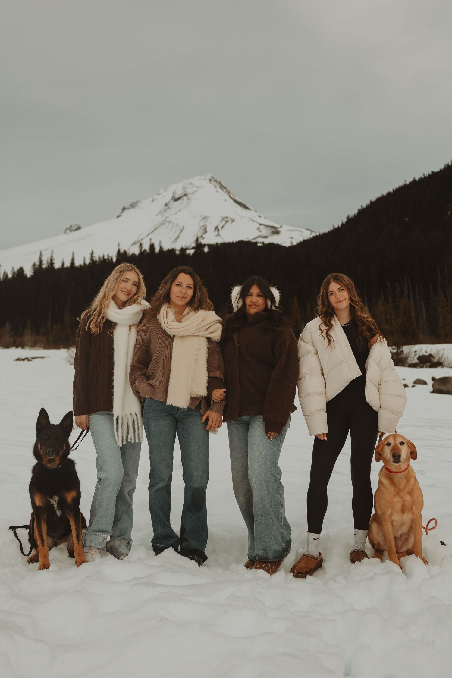 Four women with two dogs pose in winter outfits against a snowy mountain landscape.