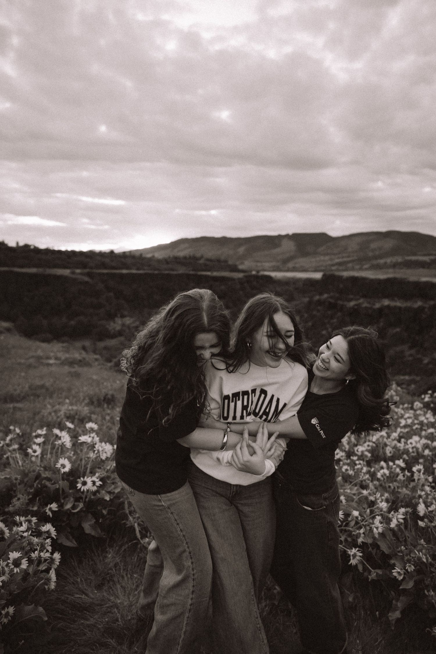 Three young women laughing and hugging in a wildflower field with rolling hills under cloudy skies, black and white photo.