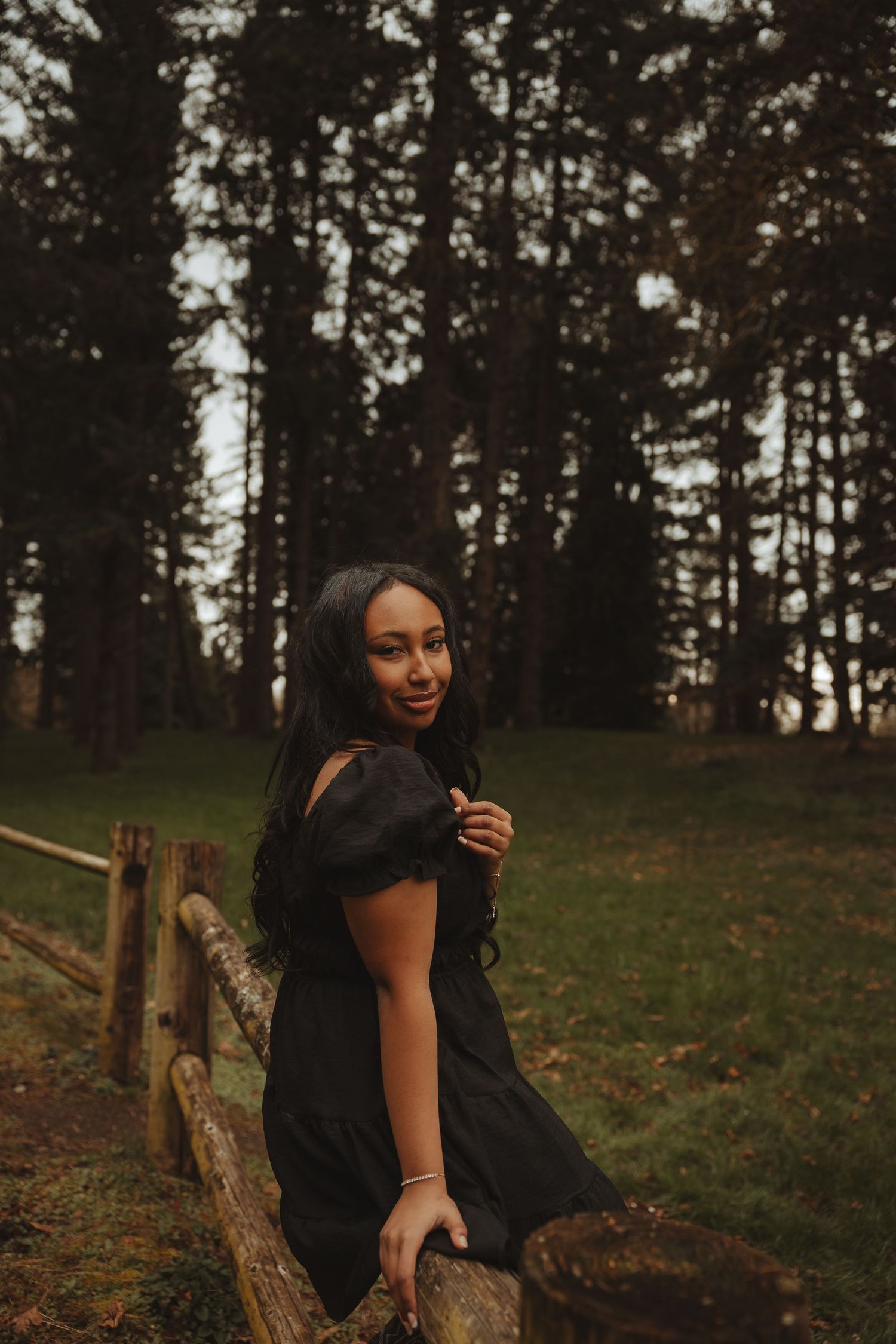 A person in a black dress poses playfully in a wooded outdoor setting during golden hour.