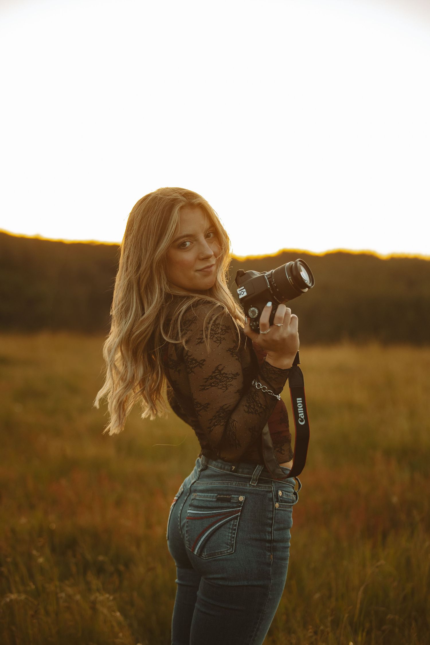 Photographer wearing lace top and jeans captures sunset in rural field.