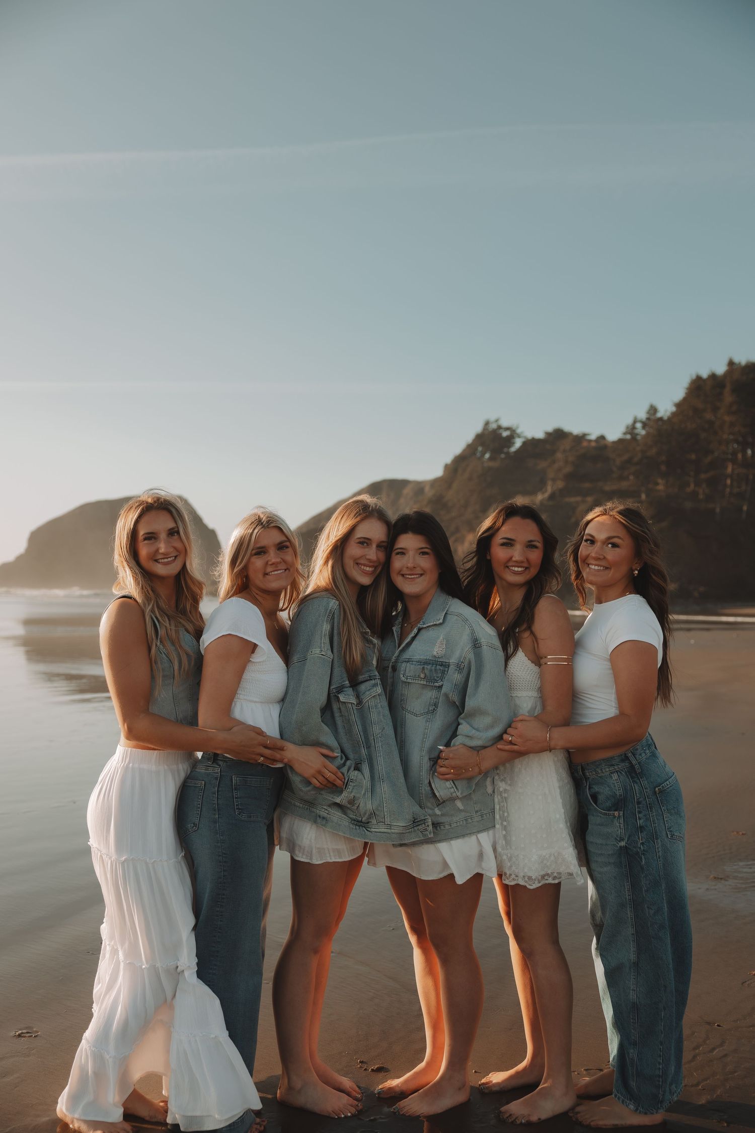 Six women posing together on a beach at golden hour, wearing white outfits and denim.