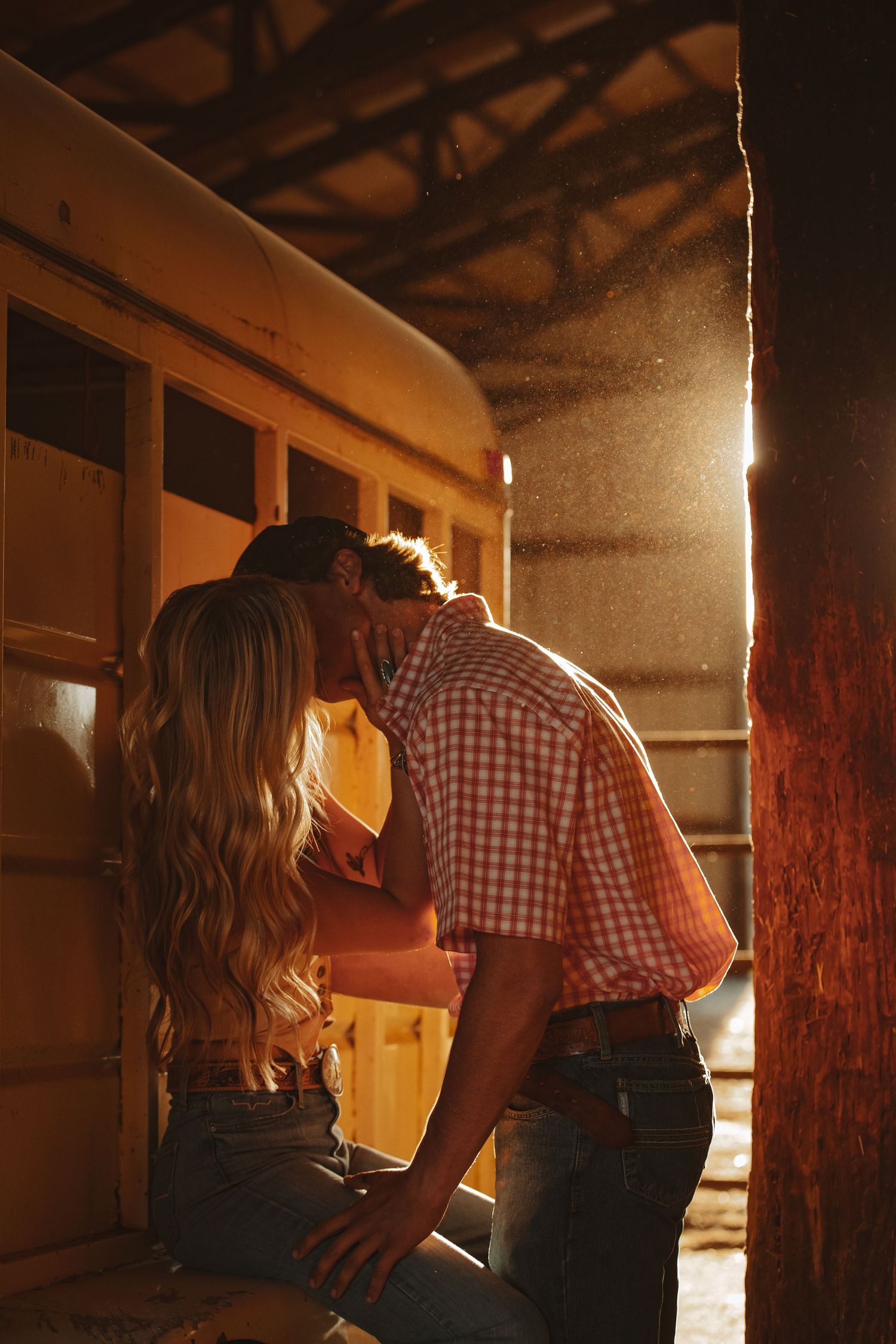 Couple sharing a romantic kiss in golden sunset light inside a rustic barn beside a vintage yellow bus.