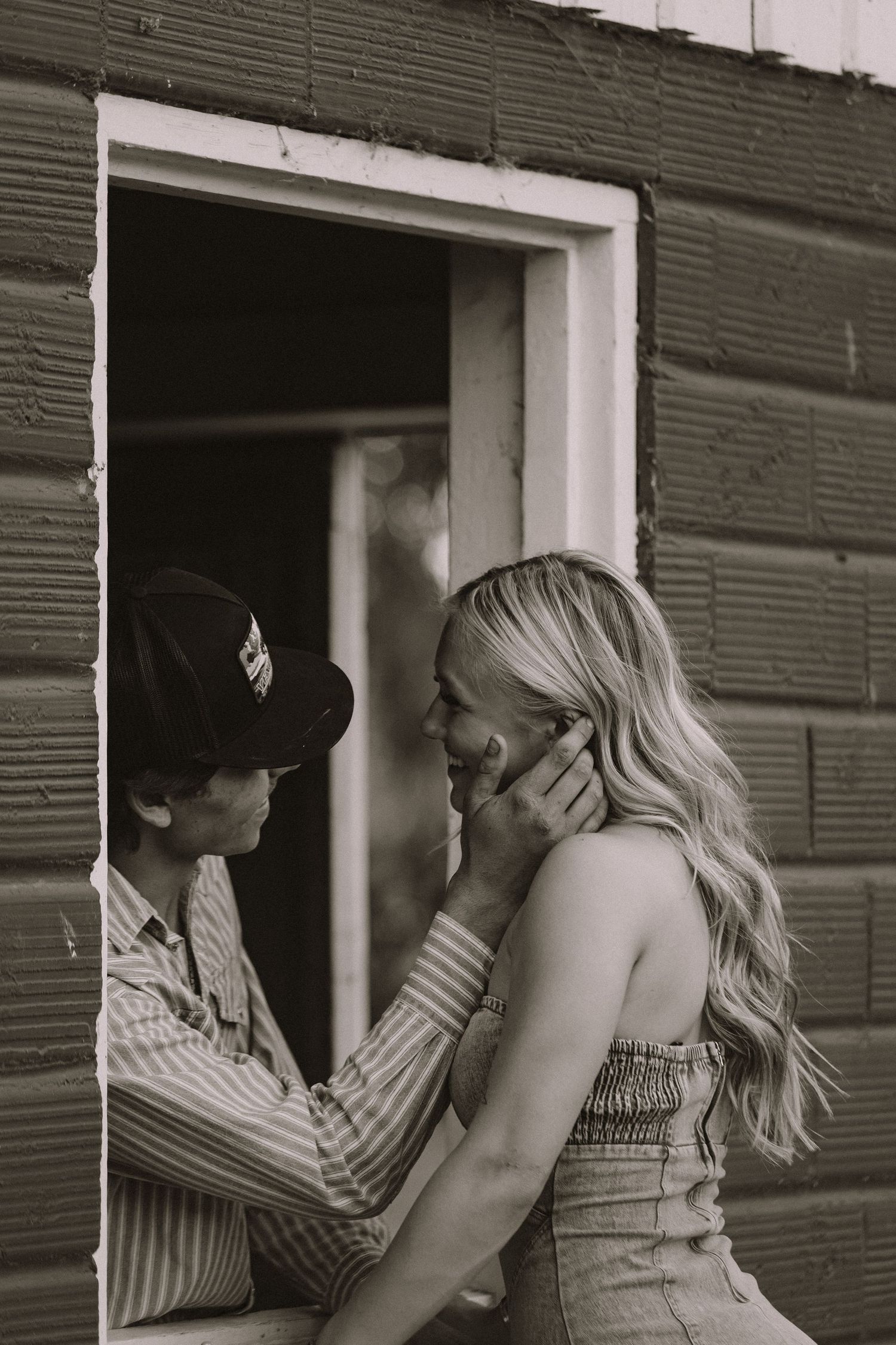 Black and white romantic photo of a man gently cupping a blonde womans face by a barn window.