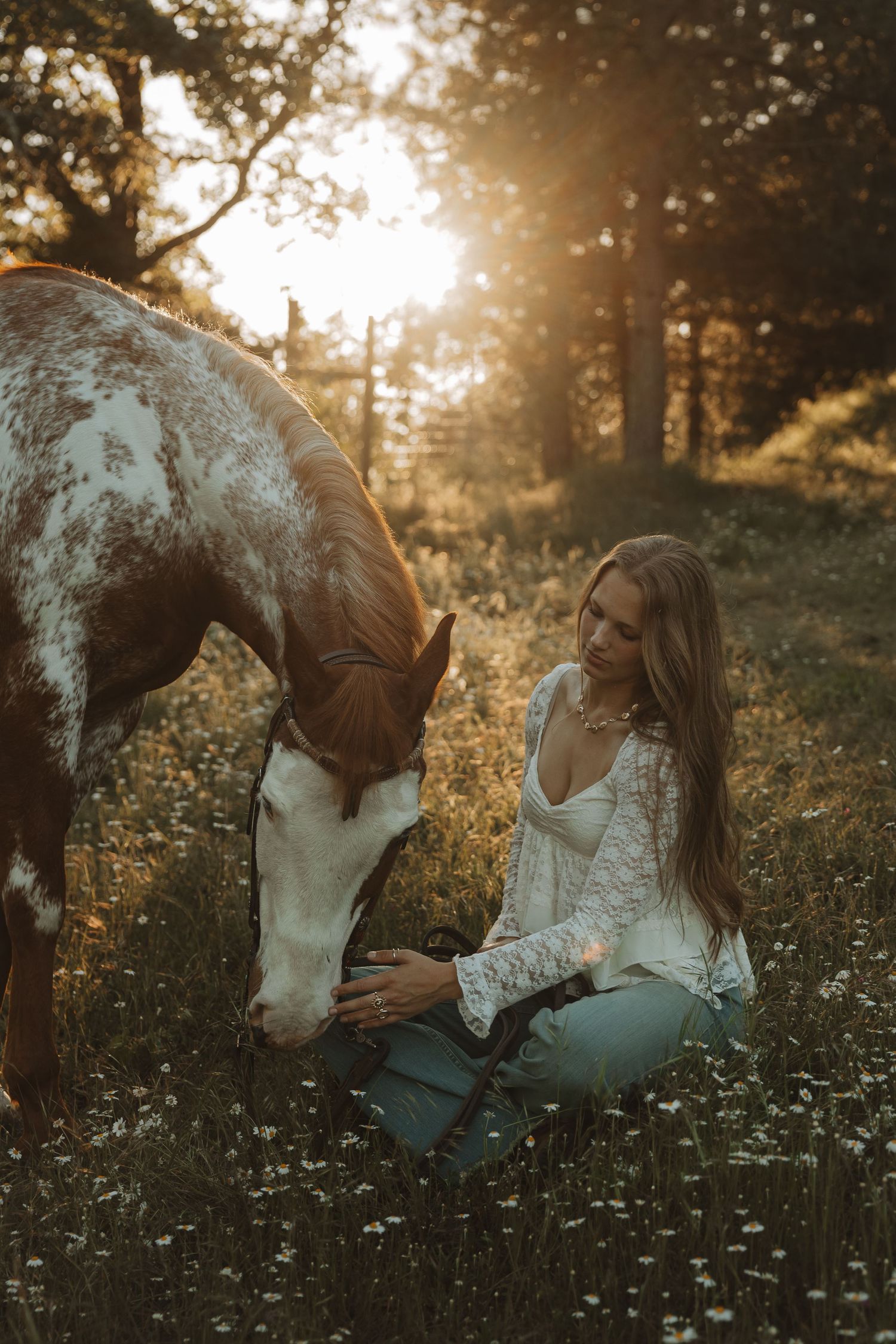 Young woman in white lace top sits in wildflower field at golden hour, gently bonding with a brown and white paint horse.
