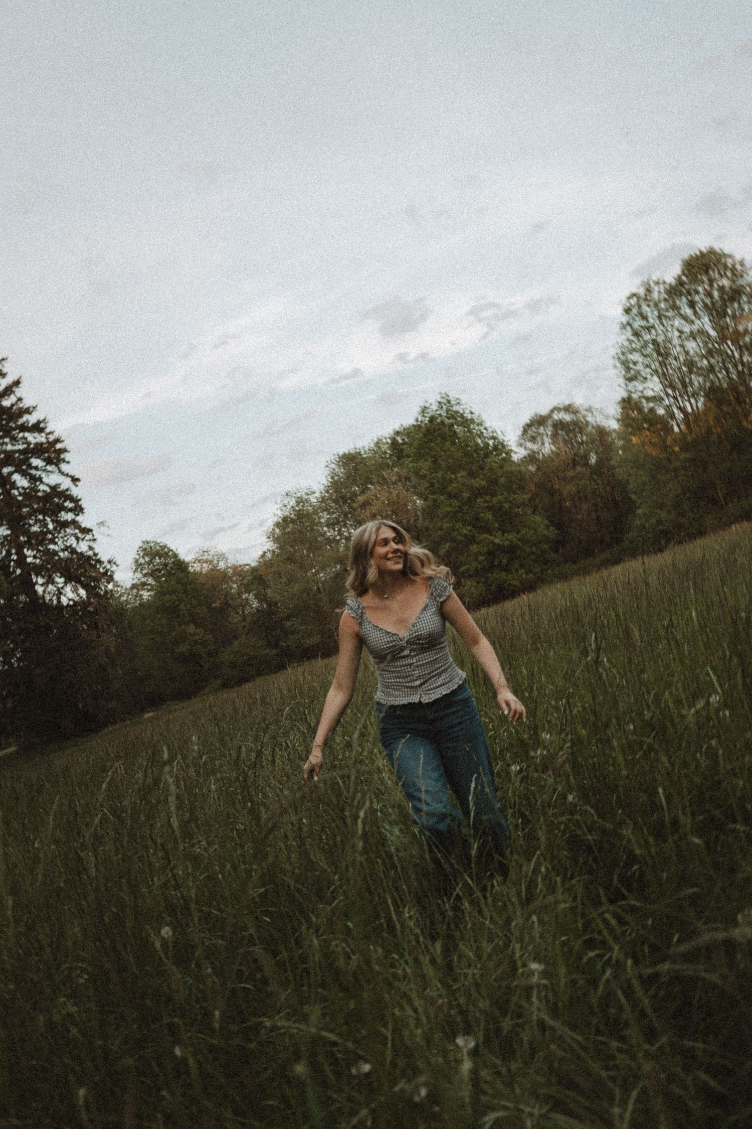 A joyful woman in a gray tank top and jeans runs through a lush green meadow with trees in the background.