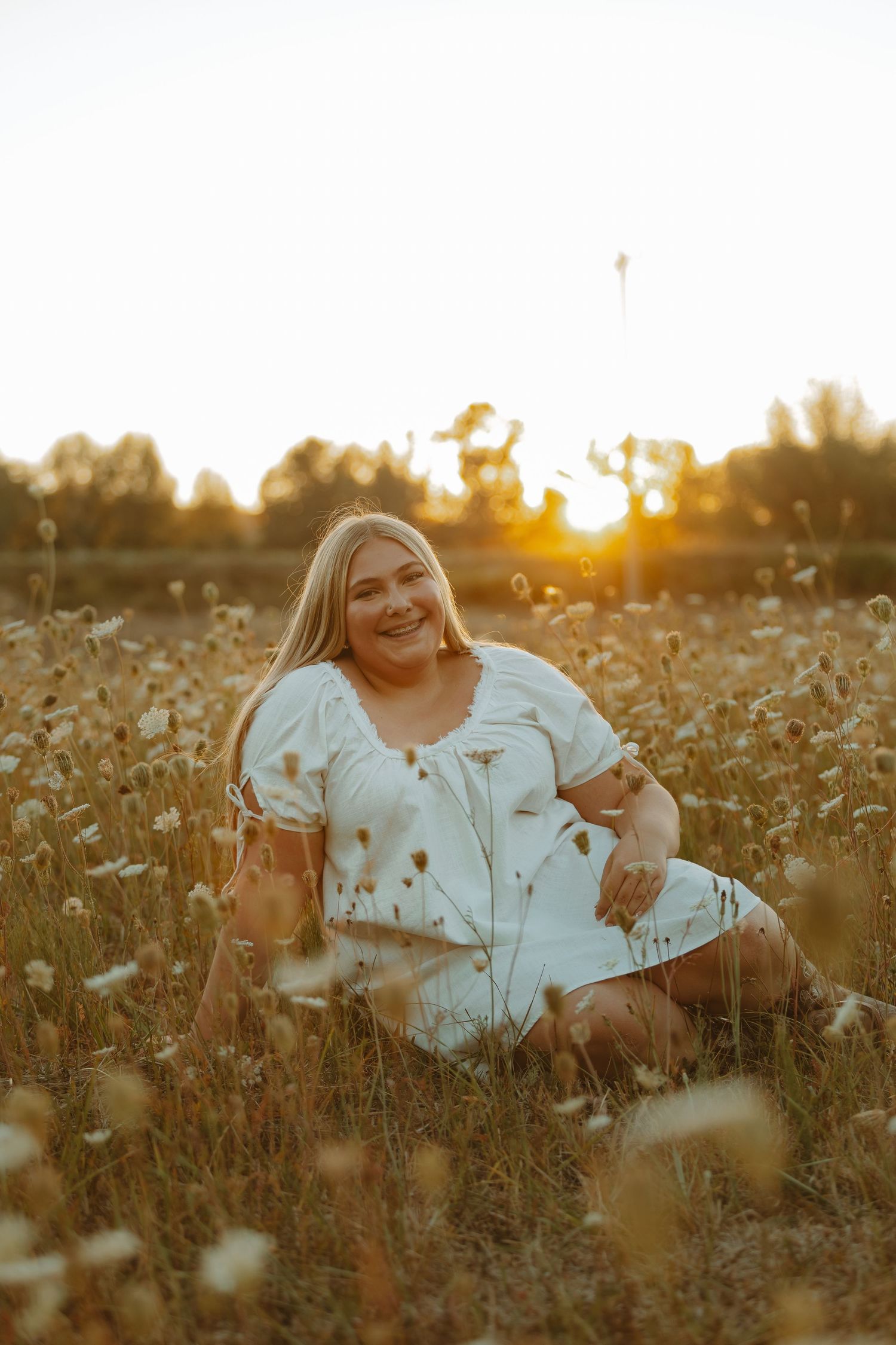 A person in a white dress sits in a golden sunlit field of wildflowers during sunset.
