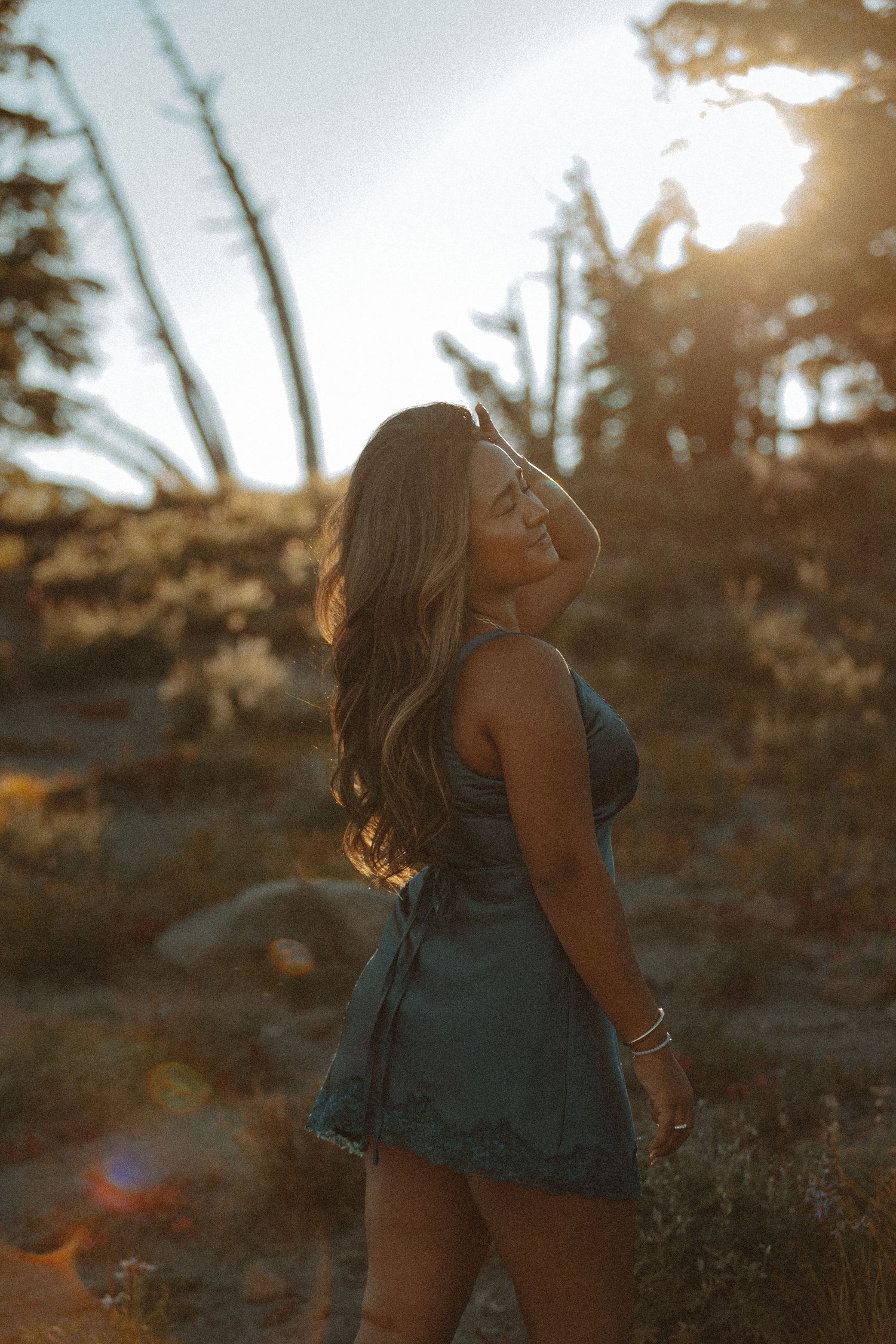 Woman in a teal dress poses joyfully against a golden sunset in a natural outdoor setting.