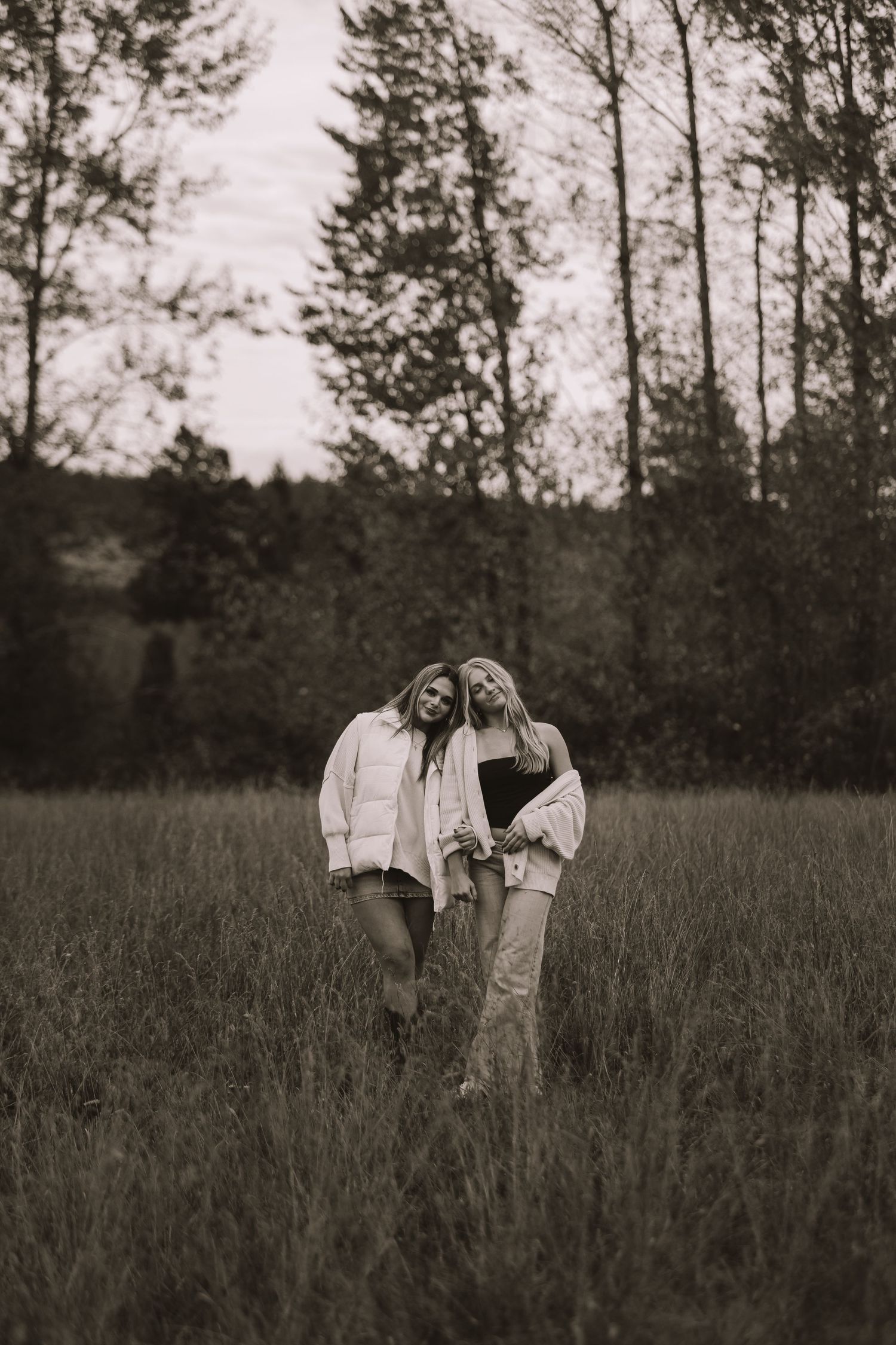 Two figures stand together in a tall grass field during golden hour with evergreen trees behind them.