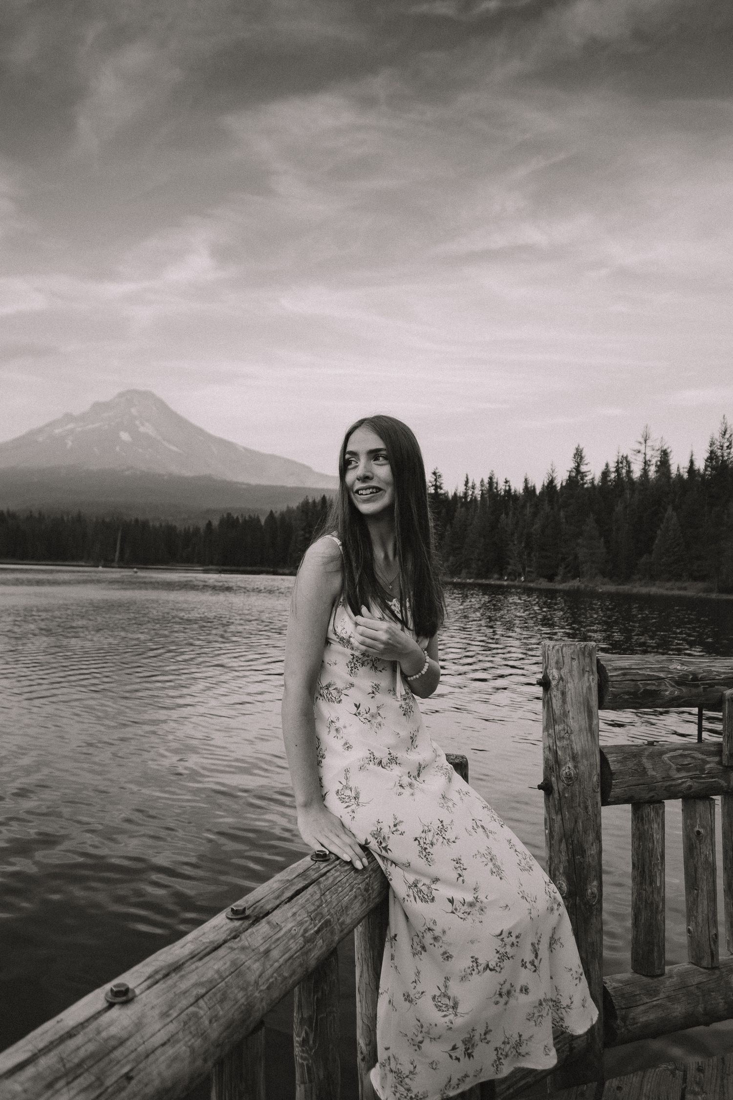 A series of black and white photos showing someone in a floral dress sitting on a wooden dock by a lake with mountains.