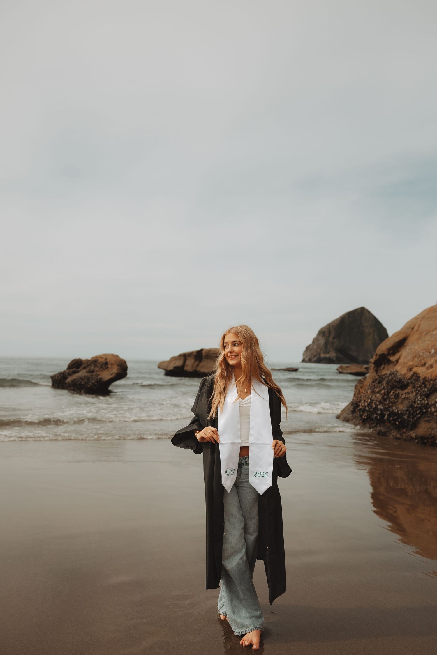 Smiling graduate in black gown and white stole stands barefoot on a sandy beach with rocky ocean backdrop.