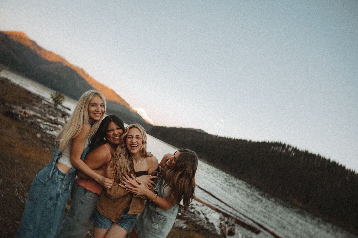 Four smiling young women hugging outdoors near mountain lake with warm sunset glow in background.
