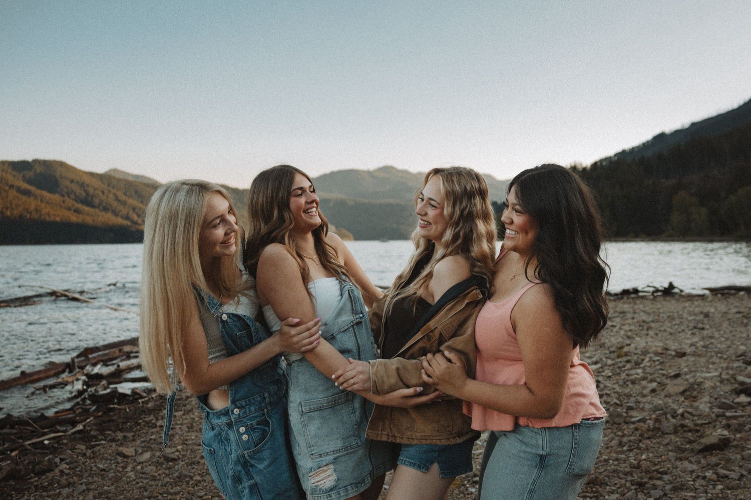 Four young women laughing and embracing on a rocky beach with a stunning mountain lake backdrop.