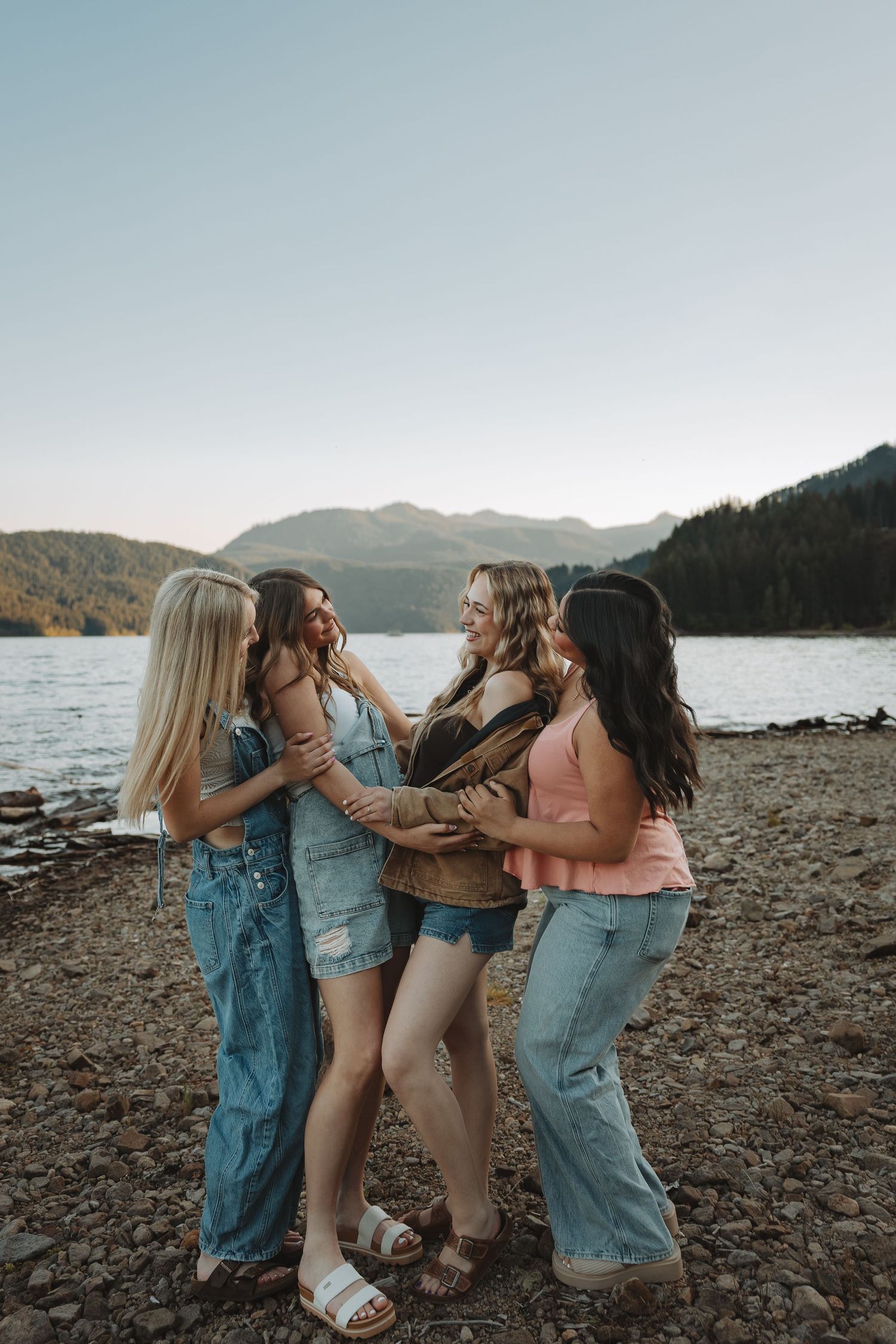 Four young women laughing and embracing on a rocky lakeshore with mountain scenery.