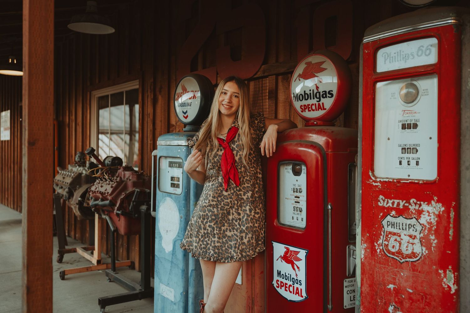 Smiling blonde woman in floral dress leans against vintage red Mobilgas Special pump at retro gas station.